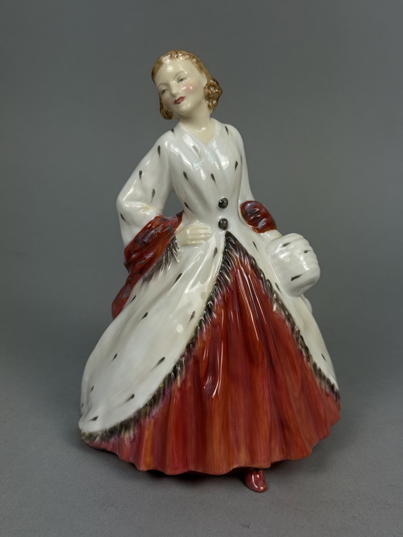Royal Doulton The Ermine Coat Figurine: This Royal Doulton porcelain figurine, titled The Ermine Coat and numbered HN 1981, depicts a standing woman in mid-20th-century attire. The figure features a vibrant red floor-length skirt, a white