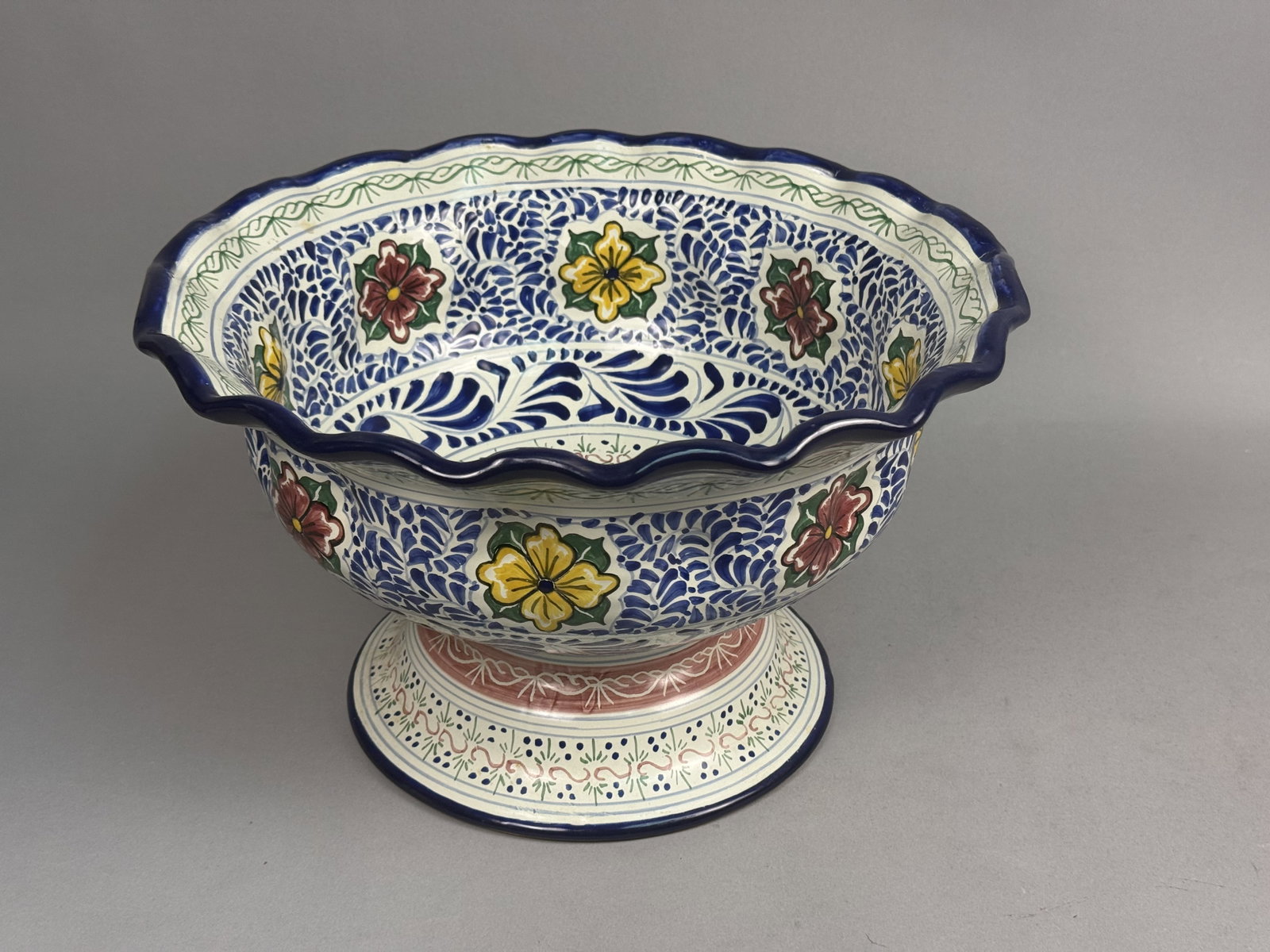 Large Mexican Talavera Footed Bowl: This hand-painted Talavera tin-glazed earthenware footed bowl originates from Puebla, Mexico. The vessel features a wide, scalloped cobalt blue rim and dense plumeado brushwork accented by large
