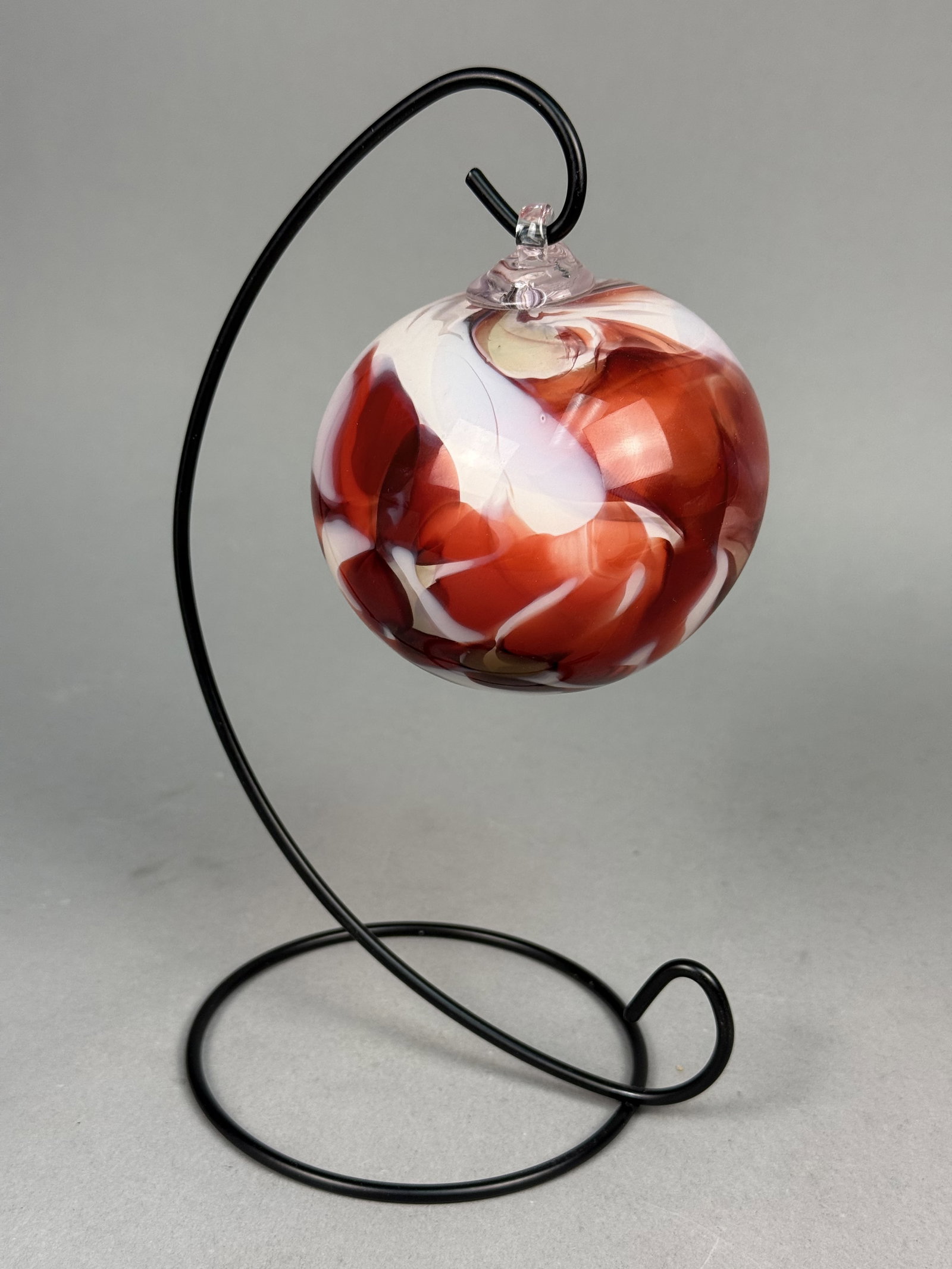 Fireworks Studios Blown Art Glass Ornament: This hand-blown spherical ornament by Fireworks Glass Studios features a dynamic swirl pattern of deep red and opaque white encased in clear glass. The piece is finished with an applied clear glass