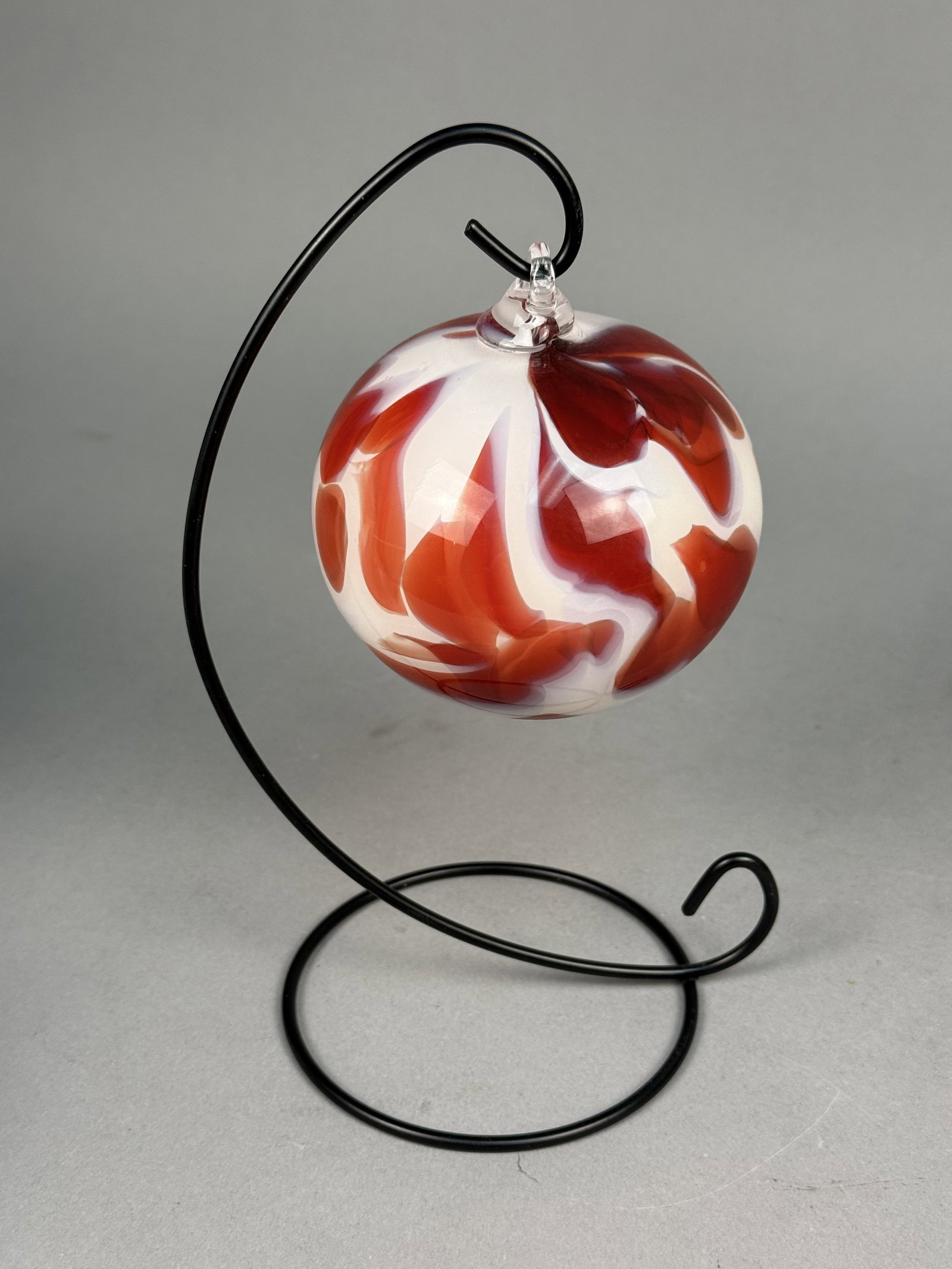 Fireworks Glass Studios Art Glass Ornament: A hand-blown glass sphere by Fireworks Glass Studios featuring an oxblood red and cream marbled swirl pattern. The piece is finished with a clear applied glass loop and is presented on a black metal