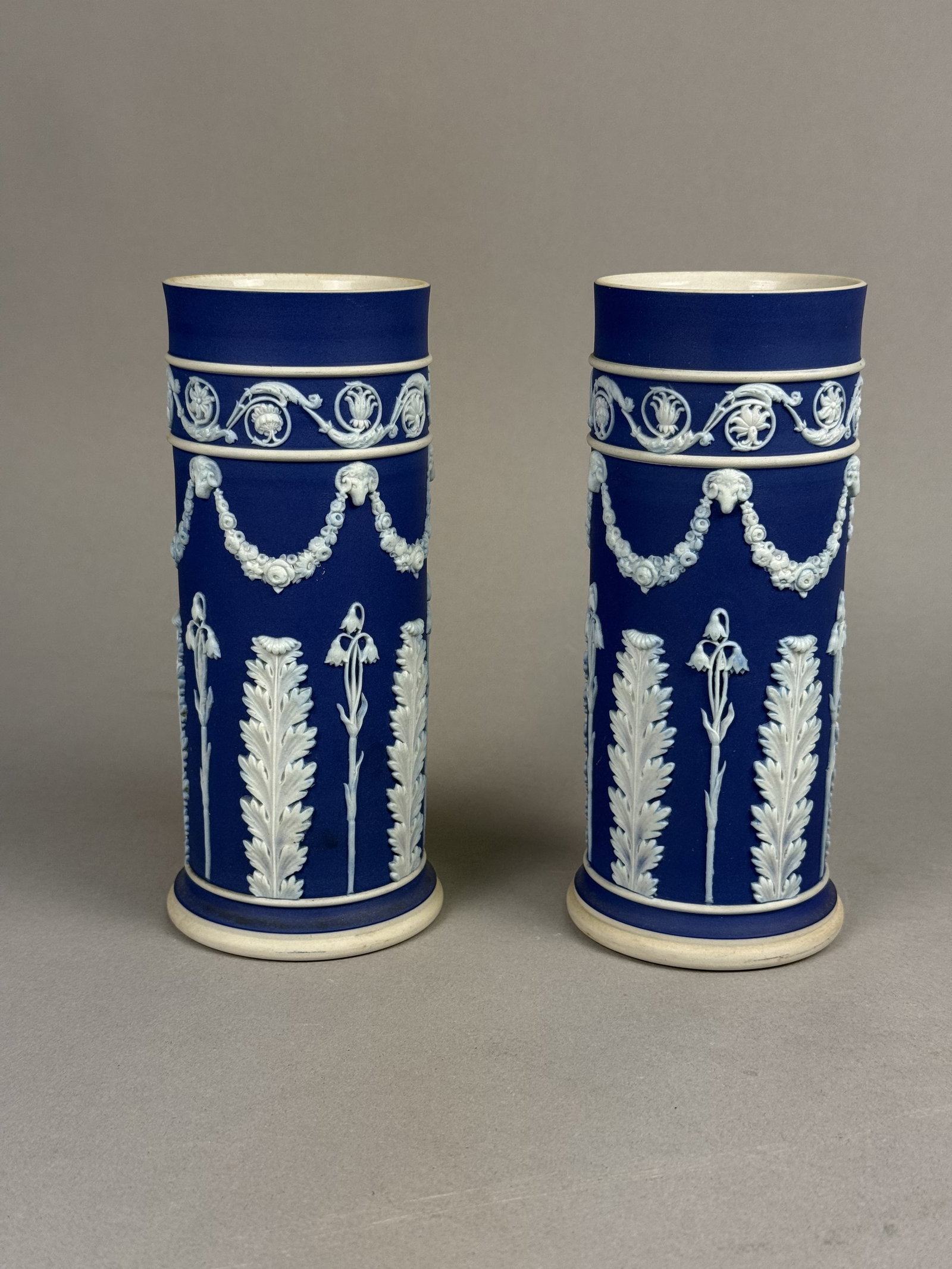 Pair of Wedgwood Jasperware Spill Vases: A pair of Wedgwood cobalt blue Jasperware spill vases featuring white Neoclassical sprigged decoration. The cylindrical forms are ornamented with a top border of scrolling foliage above rams head