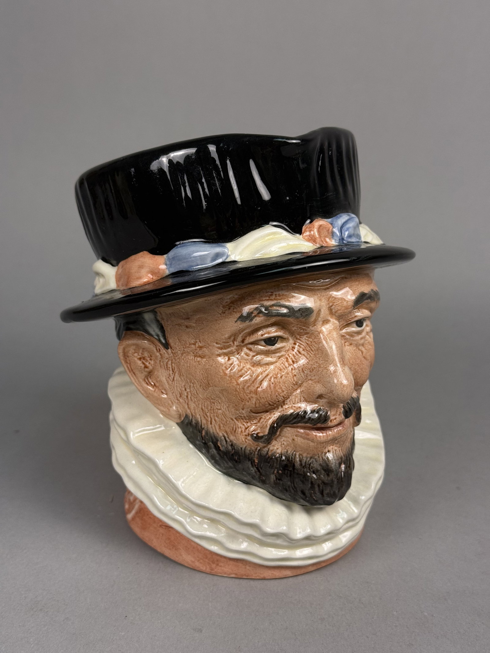 Royal Doulton Beefeater Character Jug: This Royal Doulton ceramic character jug depicts a Beefeater in traditional Tudor attire featuring a black brimmed hat and a white pleated ruff. The piece is finished in a high-gloss polychrome glaze 
