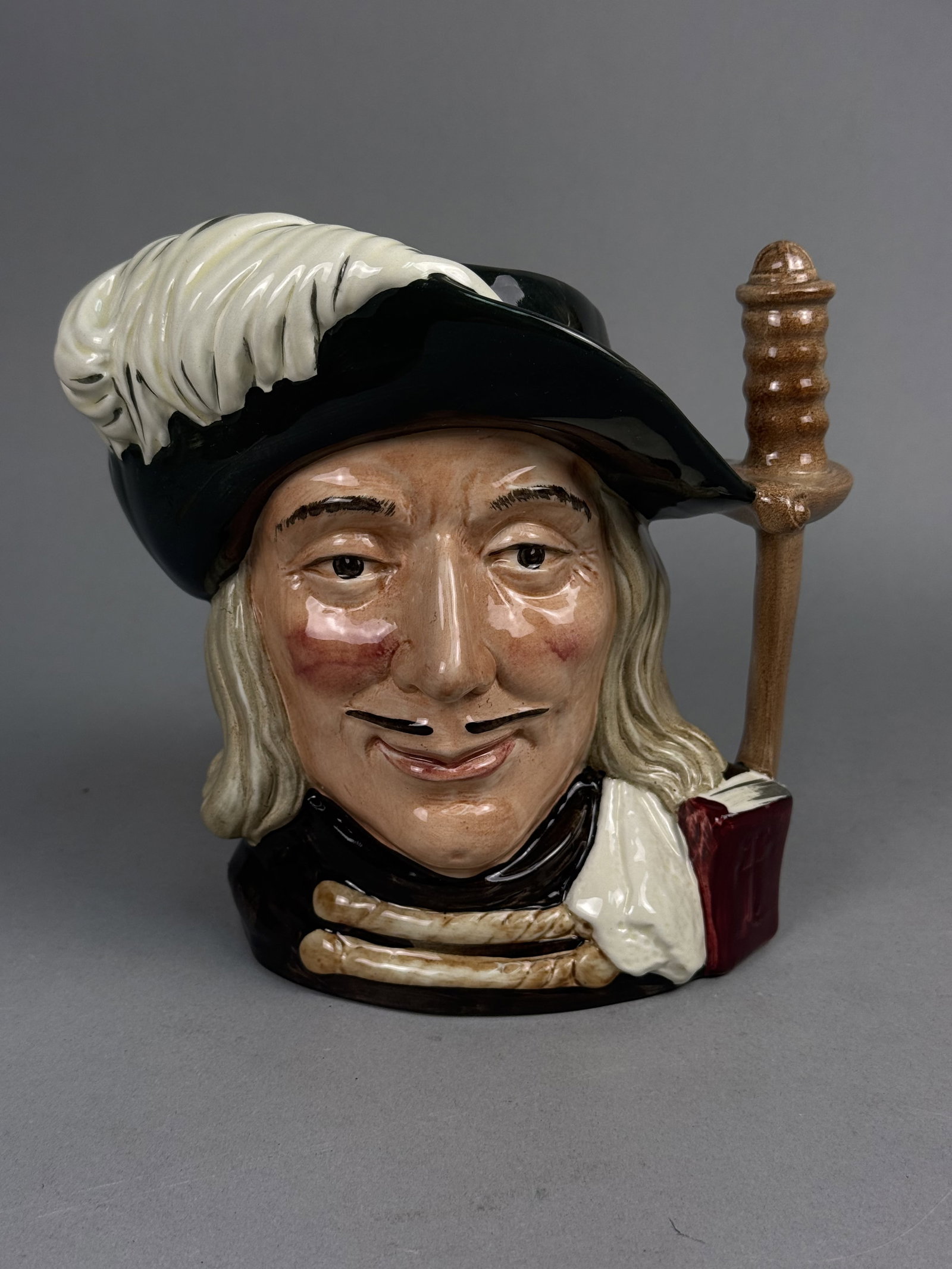 Royal Doulton Aramis Character Jug: This Royal Doulton large-scale ceramic character jug depicts the musketeer Aramis wearing a black hat with a sweeping white plume. The handle is modeled in the form of a sword hilt resting against a