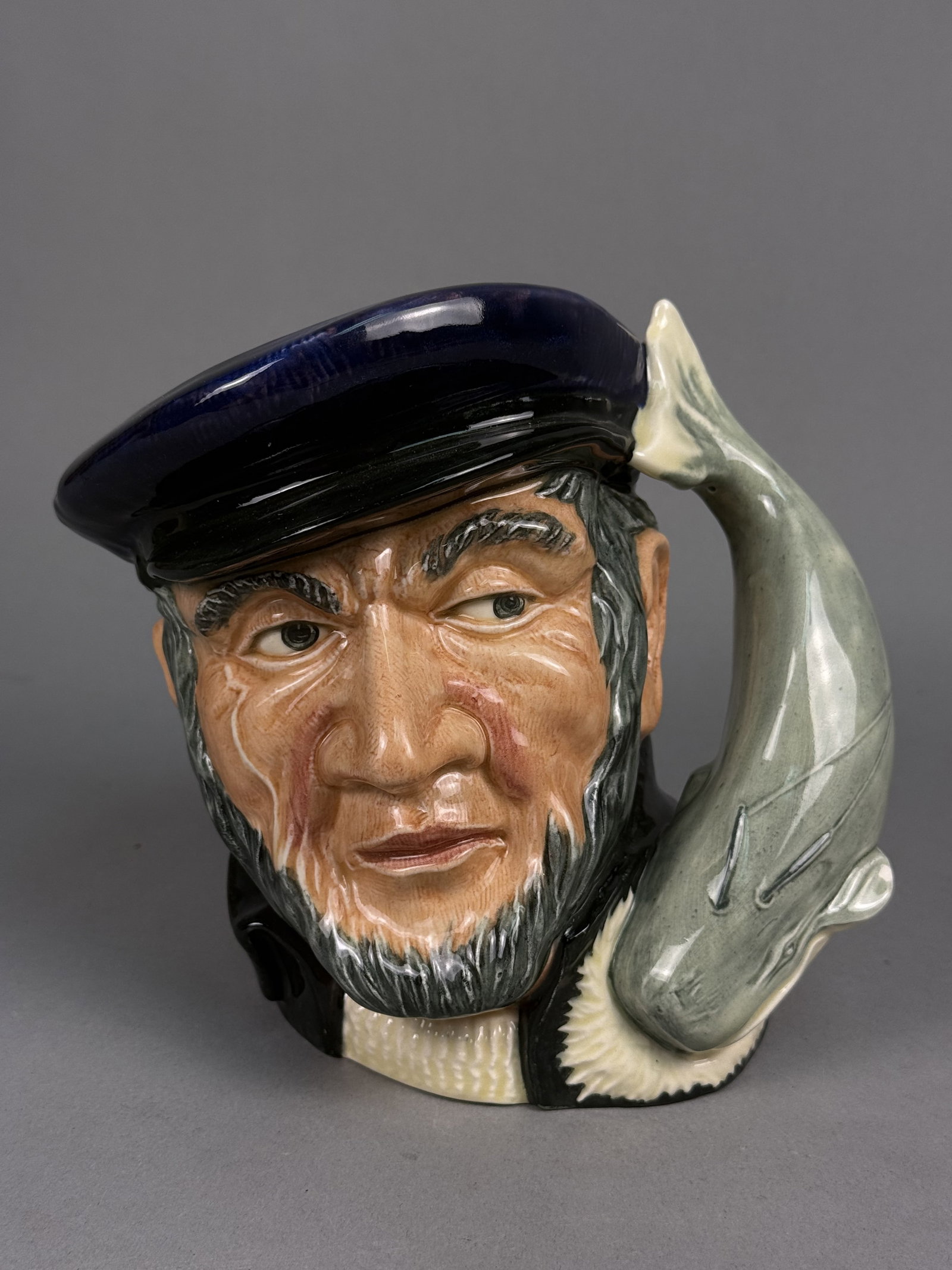 Royal Doulton Capt Ahab Character Jug: A large Royal Doulton character jug depicting Captain Ahab from Herman Melville's Moby Dick. The glazed ceramic figure features a weathered expression with a gray beard and dark blue cap, accented by