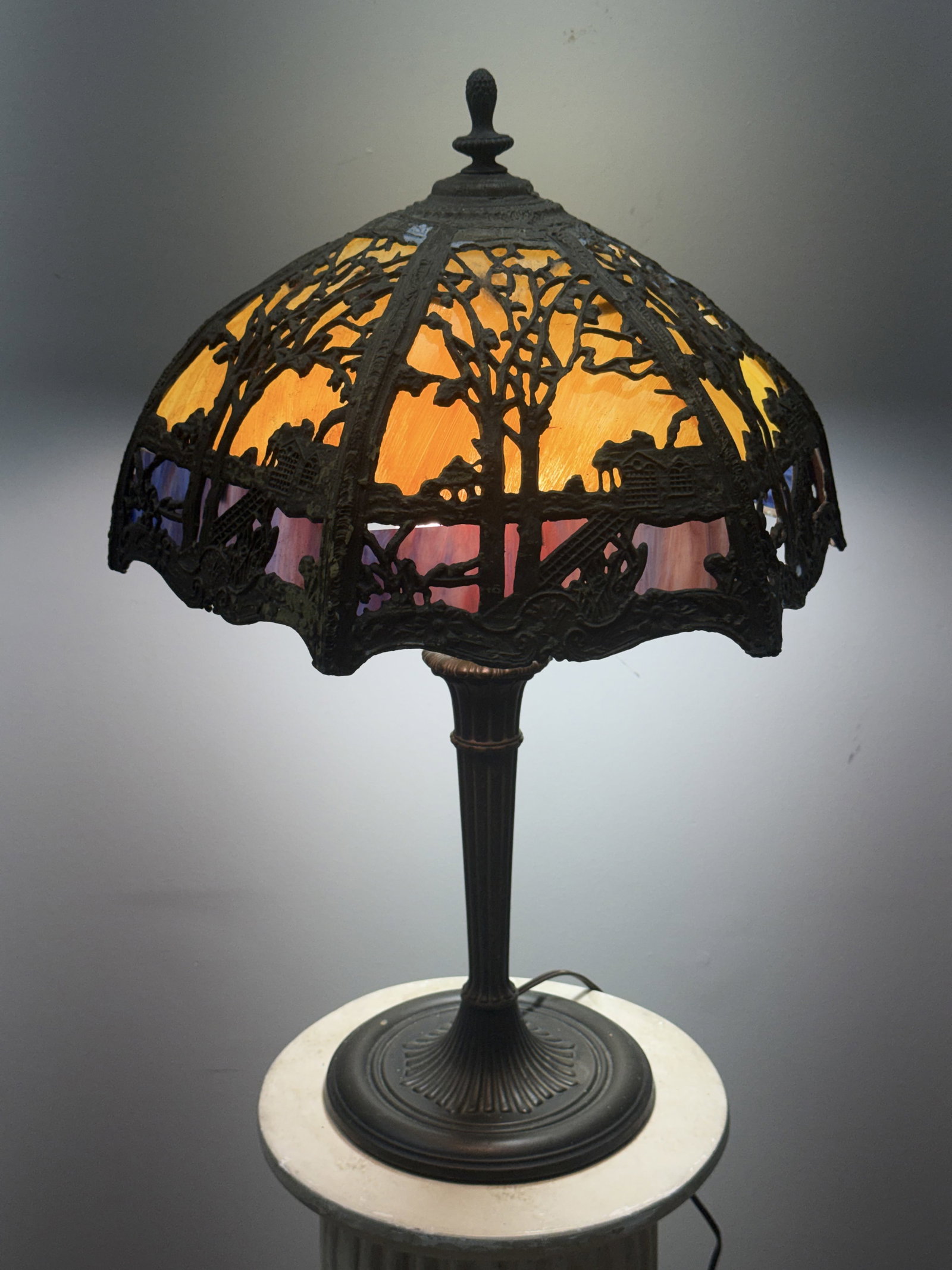 Scenic Overlay Two-Tone Slag Glass Lamp: A vintage table lamp featuring a six-panel slag glass shade with a cast metal filigree overlay depicting a continuous landscape of gnarled trees and houses. The shade uses variegated amber glass for