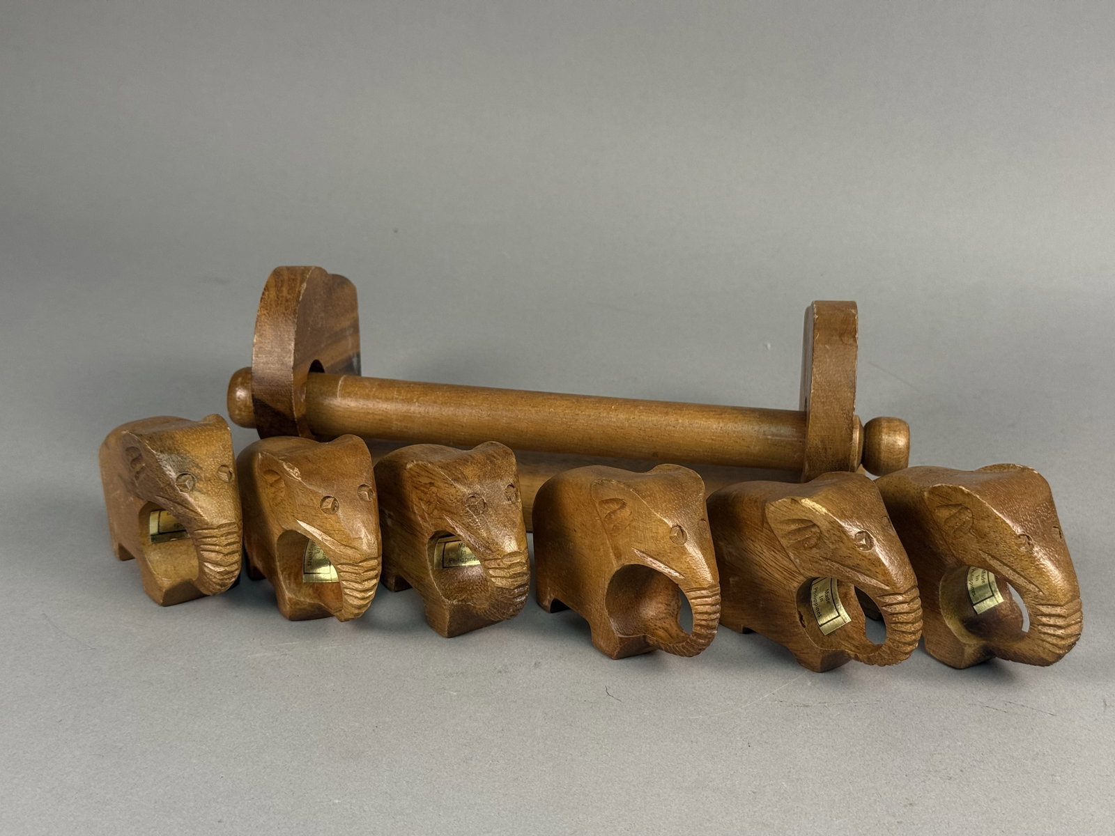 Six Carved Wood Elephant Napkin Rings: A set of six hand-carved wooden napkin rings in the form of elephants with an accompanying display stand. Each piece features detailed carving to the face and ears, with the trunk curling beneath the