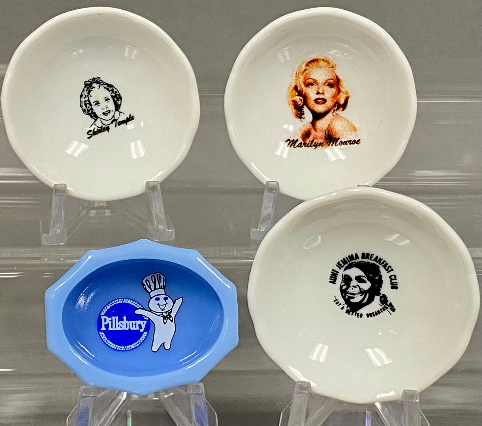 Collection of Four Miniature Advertising Dishes: A collection of four miniature dishes comprising three white glazed ceramic examples with tripod feet and one light blue opaque glass dish. Each piece features a transfer-printed design depicting