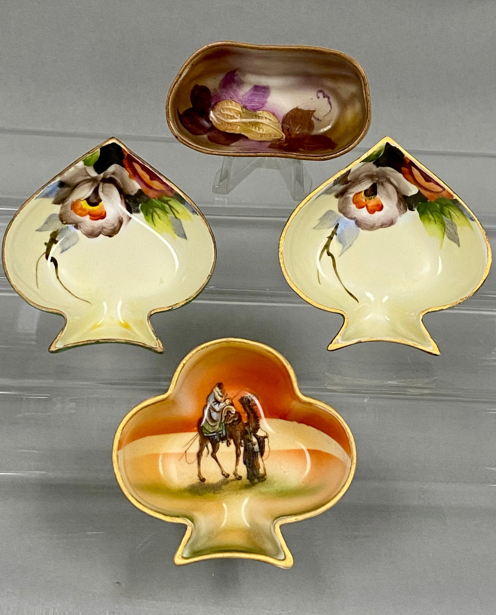 Collection of Four Noritake Dishes: A collection of four hand-painted porcelain dishes by Noritake featuring card suit and novelty peanut shapes. The assortment includes two spade-shaped floral dishes, a club-shaped dish with a desert s