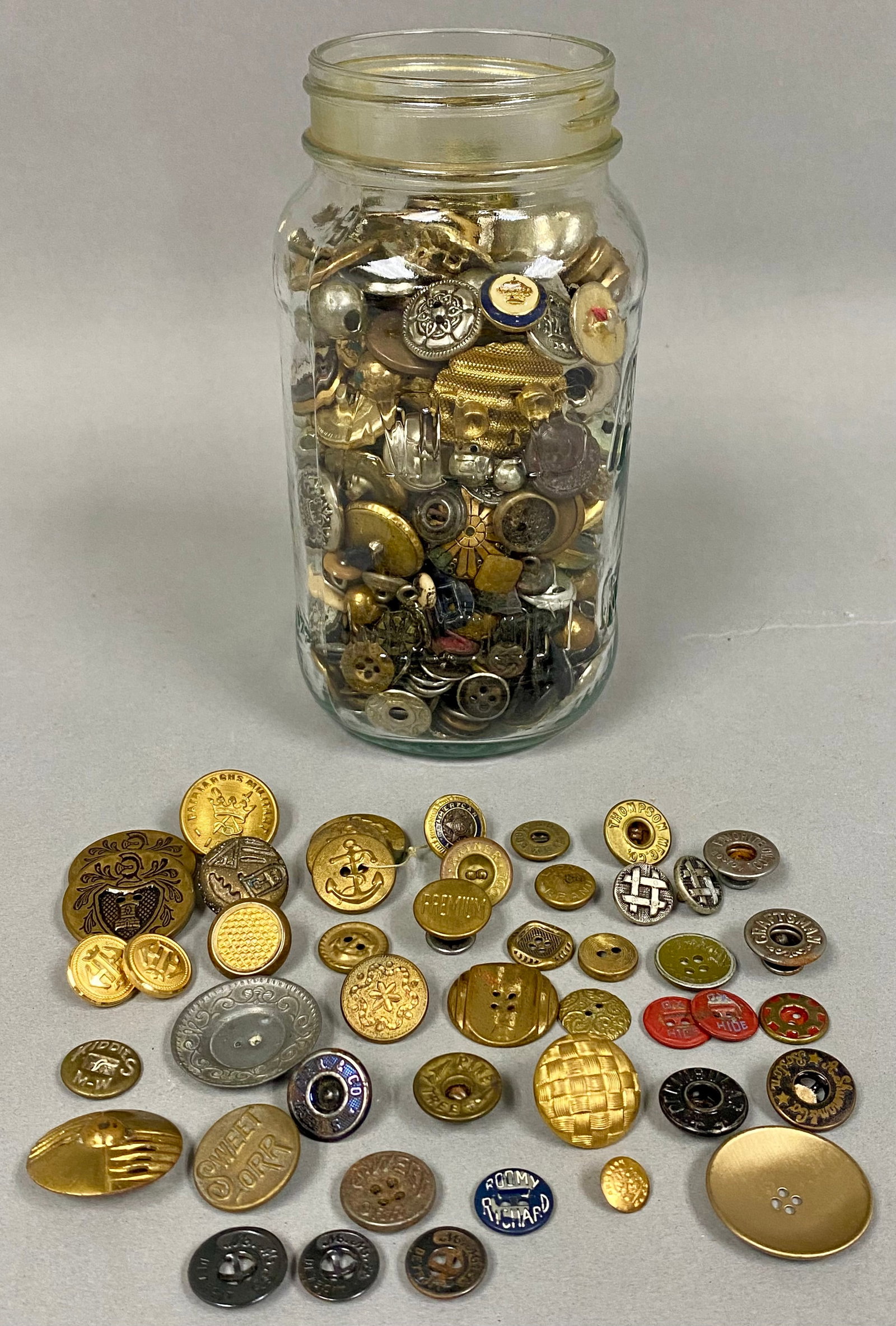 Antique Metal Workwear and Military Buttons: A diverse collection of vintage and antique metal buttons, primarily constructed from brass and copper, housed within a clear glass jar. Specimens include notable workwear brands such as Sweet-Orr, Ca