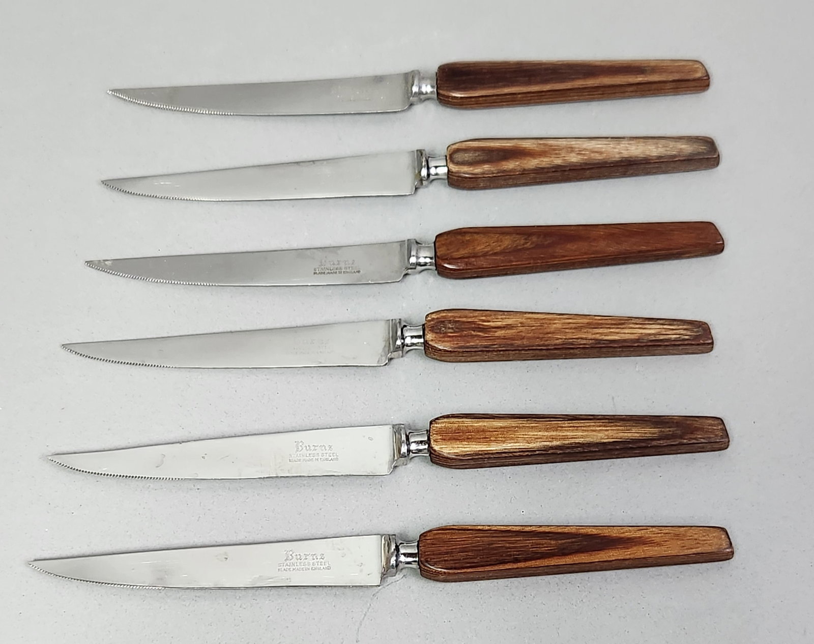 Six Burns Steak Knives: A set of six Burns steak knives featuring serrated stainless steel blades and contoured dark hardwood handles. Total length: 8.75 inches Blade length: 4.75 inches. The set shows signs of regular use,