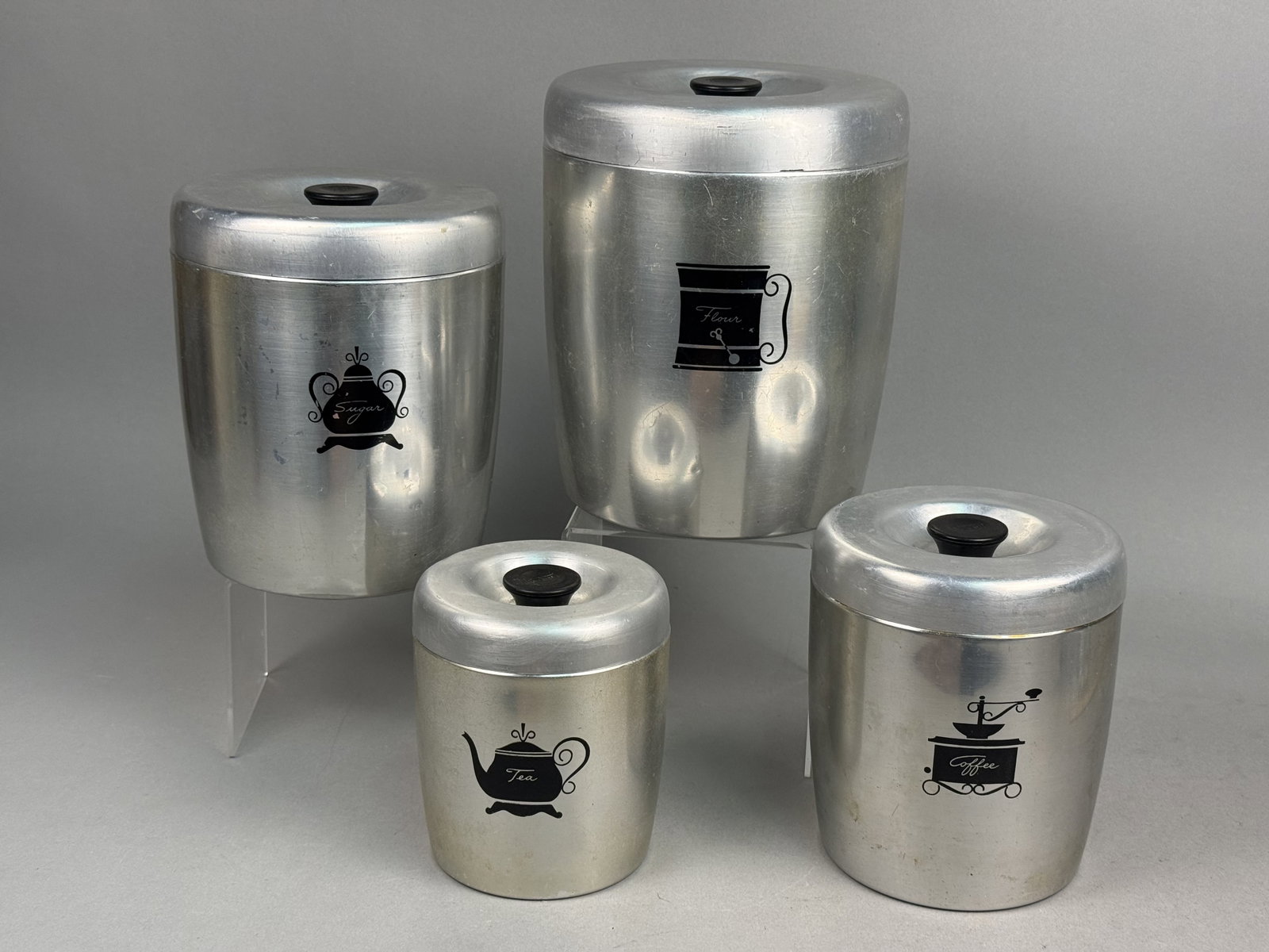 West Bend Mid-Century Aluminum Canisters: A set of four graduated nesting kitchen canisters manufactured by West Bend. Each is constructed of spun aluminum with black plastic lid knobs and features black stamped graphic illustrations for flou