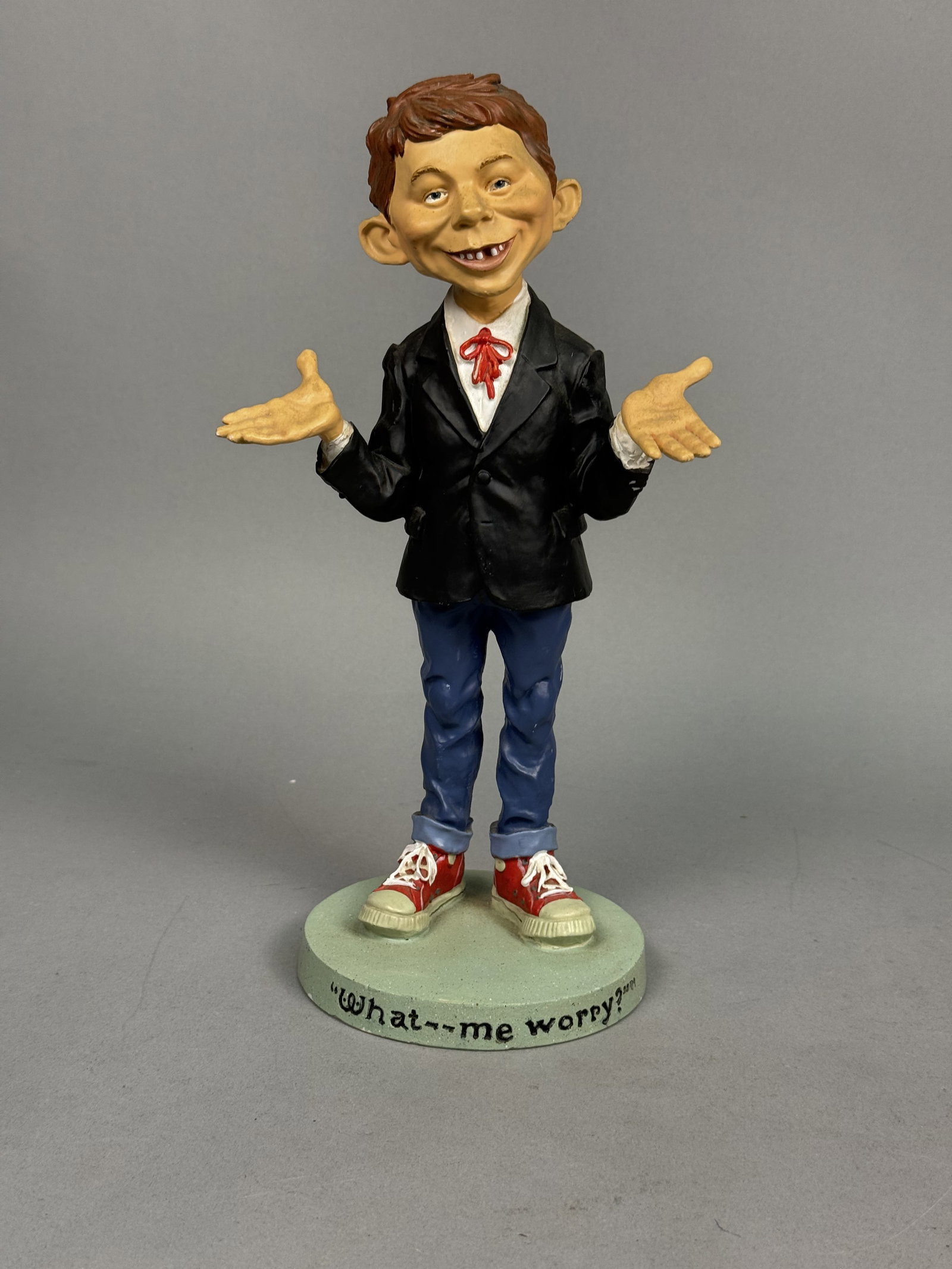 MAD Magazine Alfred E. Neuman Figurine: This licensed hand-painted figurine depicts the iconic MAD Magazine mascot, Alfred E. Neuman, in his signature shrugging pose atop a circular sage green base. The character is dressed in a black