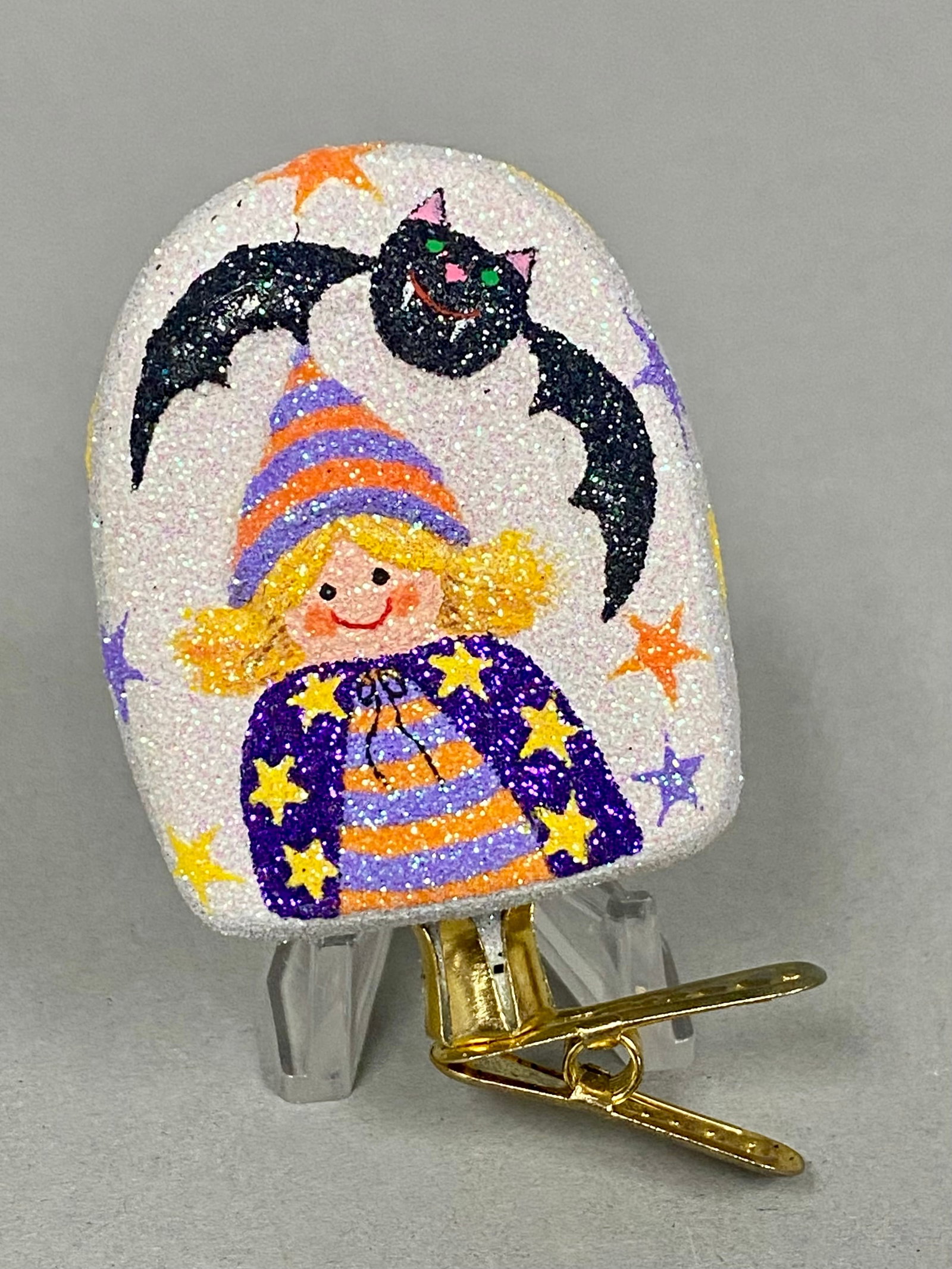 Vintage Halloween Witch Tombstone Clip: This double-sided Halloween clip-on ornament features a tombstone-shaped body with an iridescent white glitter finish. One side depicts a stylized young witch in a star-patterned cape and striped