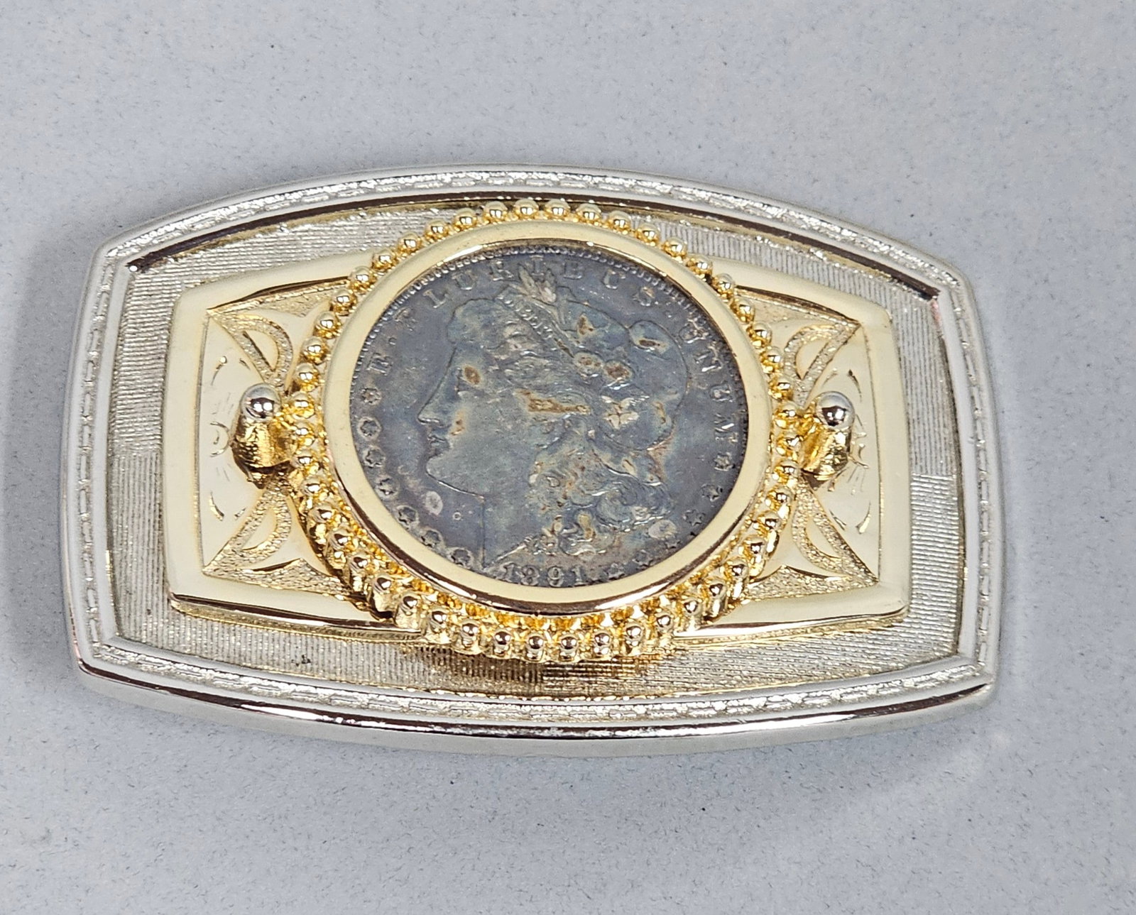 1891 Morgan Silver Dollar Belt Buckle: This rectangular western-style belt buckle features a center-set authentic United States 1891 Morgan Silver Dollar. The coin is mounted within a raised gold-tone beaded bezel on an engraved plate,