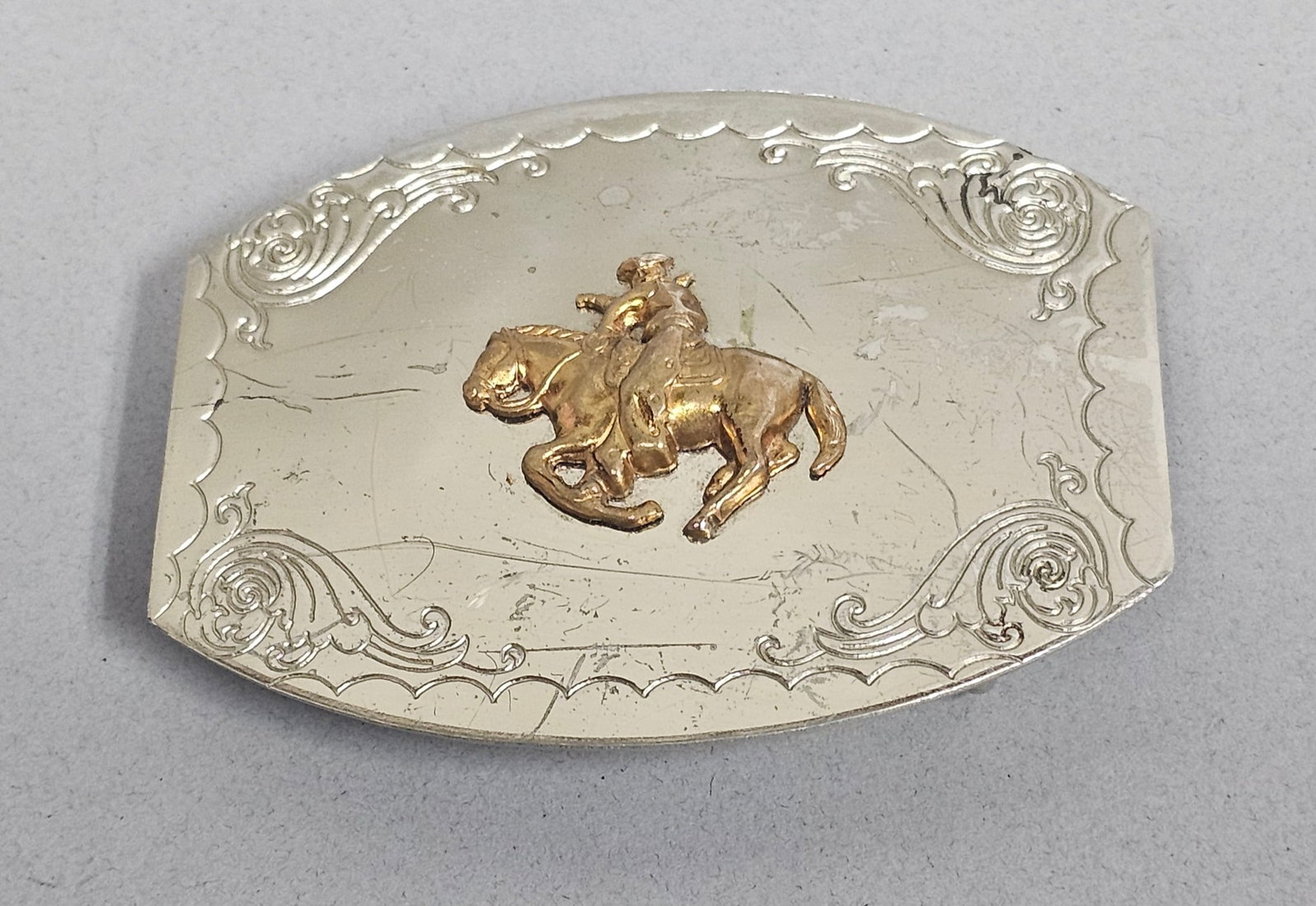 Nickel Silver Bucking Bronco Buckle: A mid-century Western-style belt buckle featuring a raised gold-tone relief of a cowboy on a bucking bronco, set against an engraved nickel silver plate. The buckle is constructed with scalloped