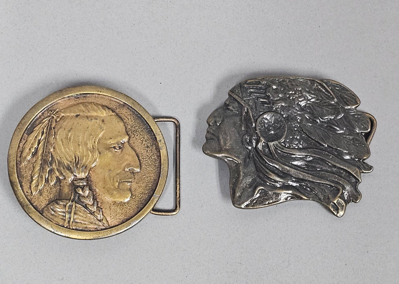 Two Vintage Native American Belt Buckles: Two vintage cast metal belt buckles featuring Native American profile motifs. One circular brass-toned buckle is marked for Indiana Metal Craft and features a profile portrait with braided hair