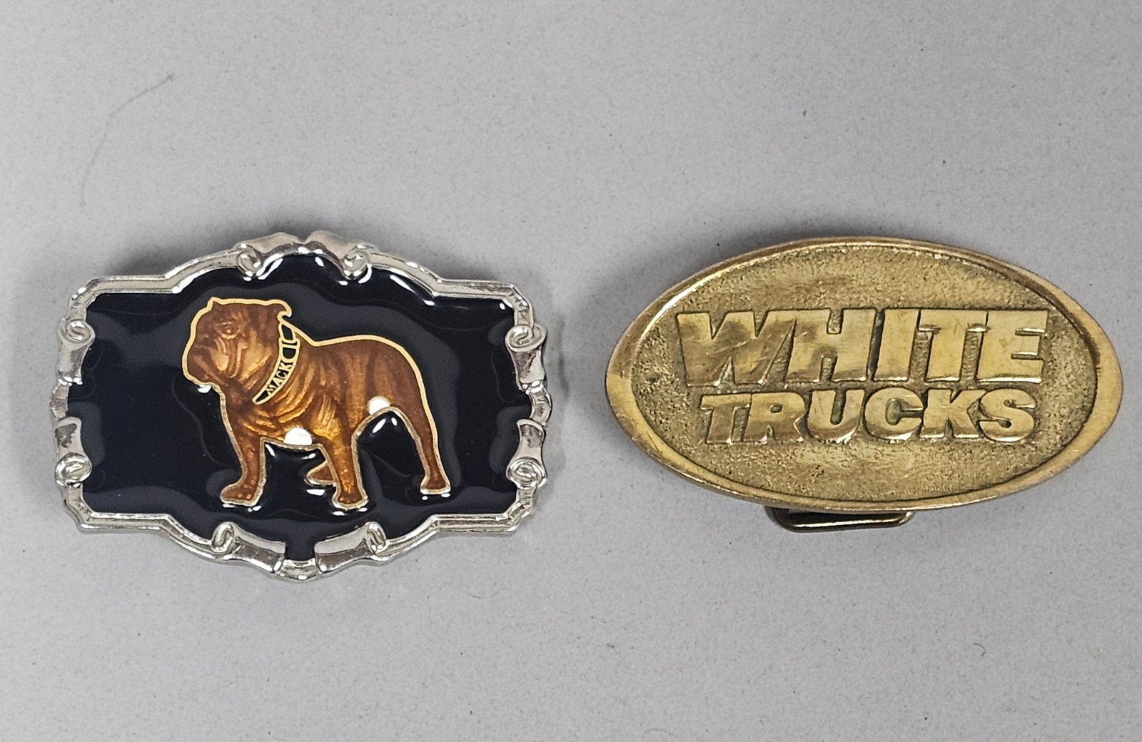 Mack and White Trucks Belt Buckles: A pair of vintage belt buckles featuring Mack and White Trucks company branding. The Mack buckle is composed of silver-toned metal with black and variegated brown enamel detailing, depicting the