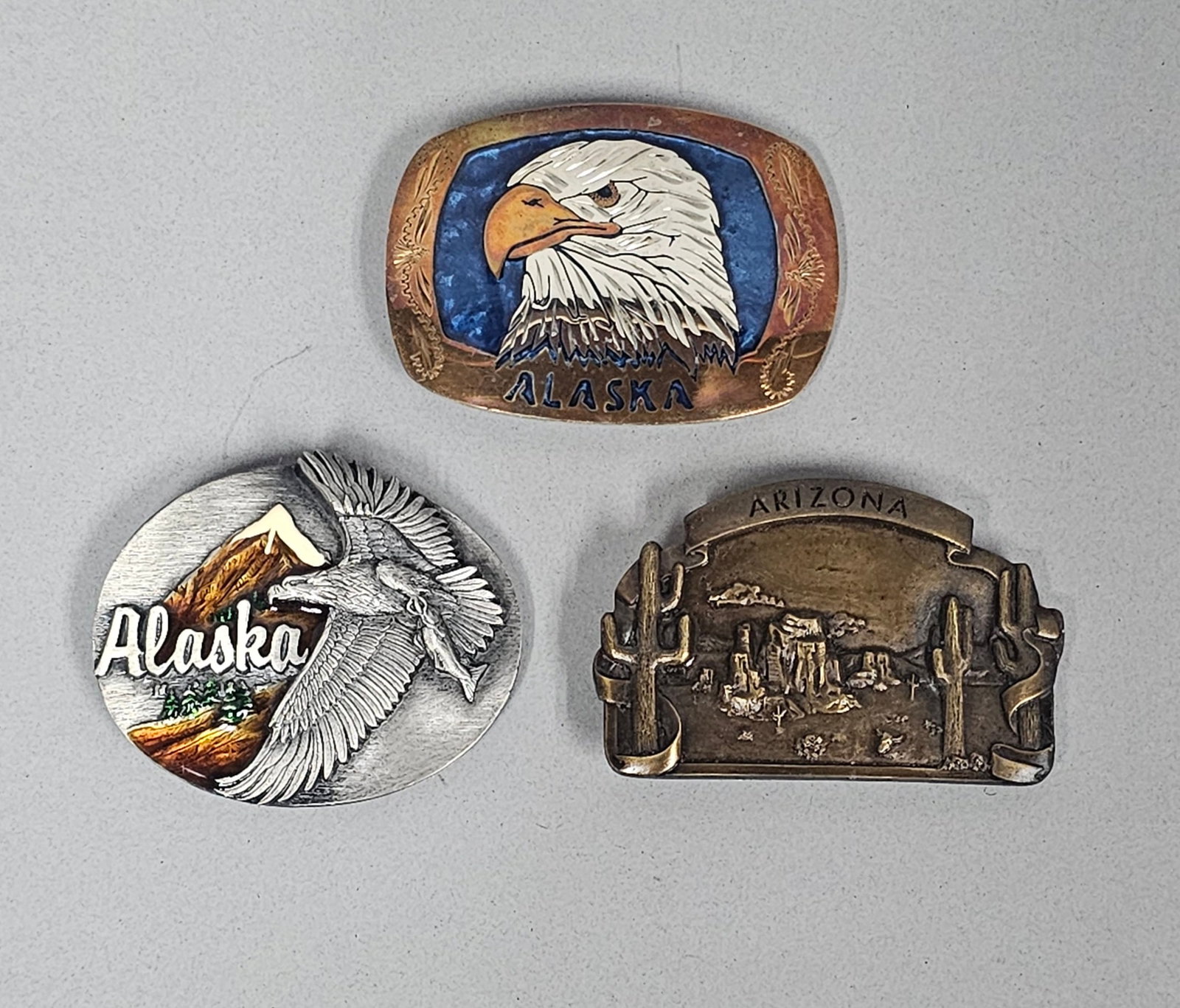 Trio of Souvenir Belt Buckles: This collection includes three commemorative belt buckles representing Alaska and Arizona, with specific examples from Bergamot Brass Works and Siskiyou Buckle Co. The designs feature high-relief