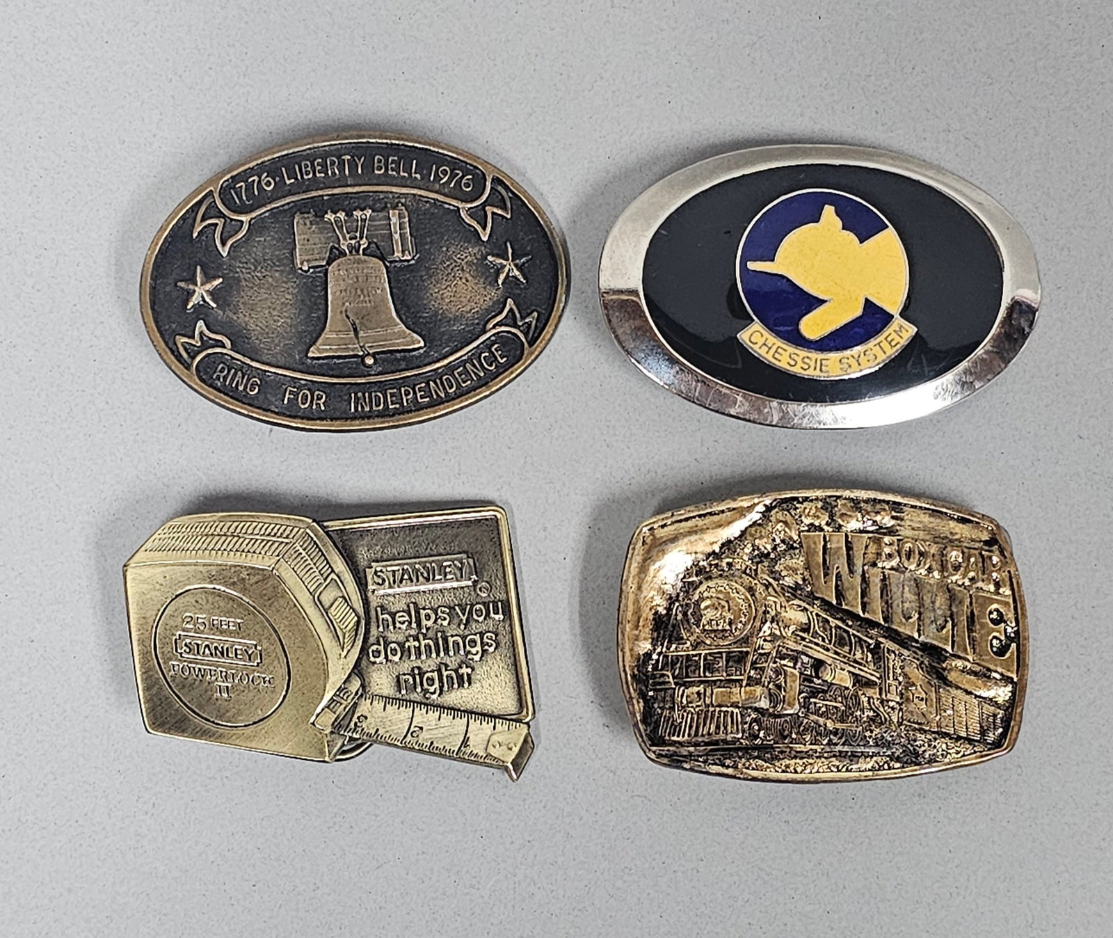 A Collection of Four Belt Buckles: A collection of four cast metal belt buckles featuring American industrial, historical, and pop-culture motifs. The assortment includes a 1776-1976 Bicentennial Liberty Bell buckle, an enameled