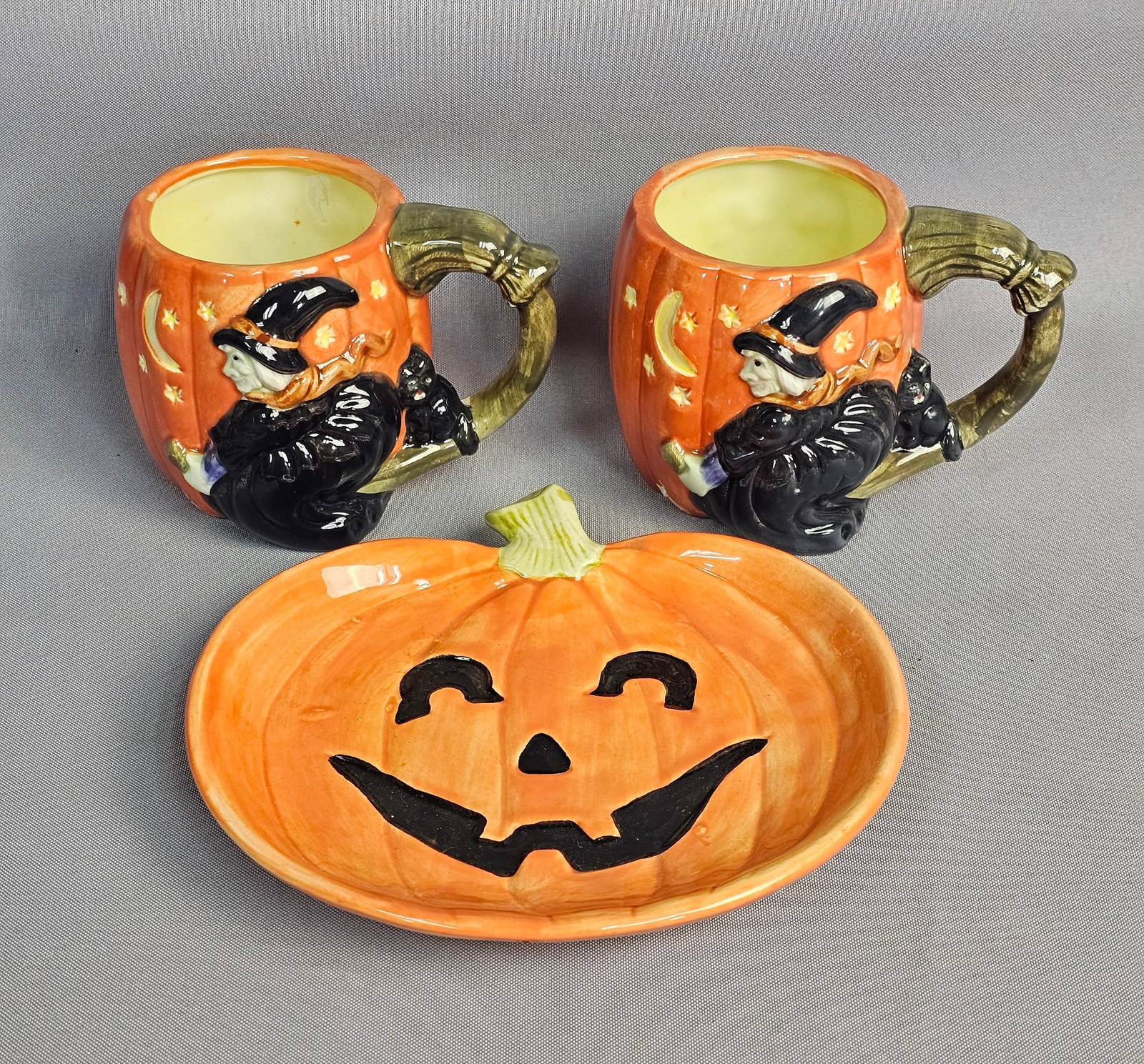 Trio of Fitz and Floyd Halloween Ceramics: This three-piece Fitz and Floyd Halloween ceramic set includes two mugs and one pumpkin-shaped serving dish featuring molded relief designs and hand-painted details. Each mug depicts a flying witch