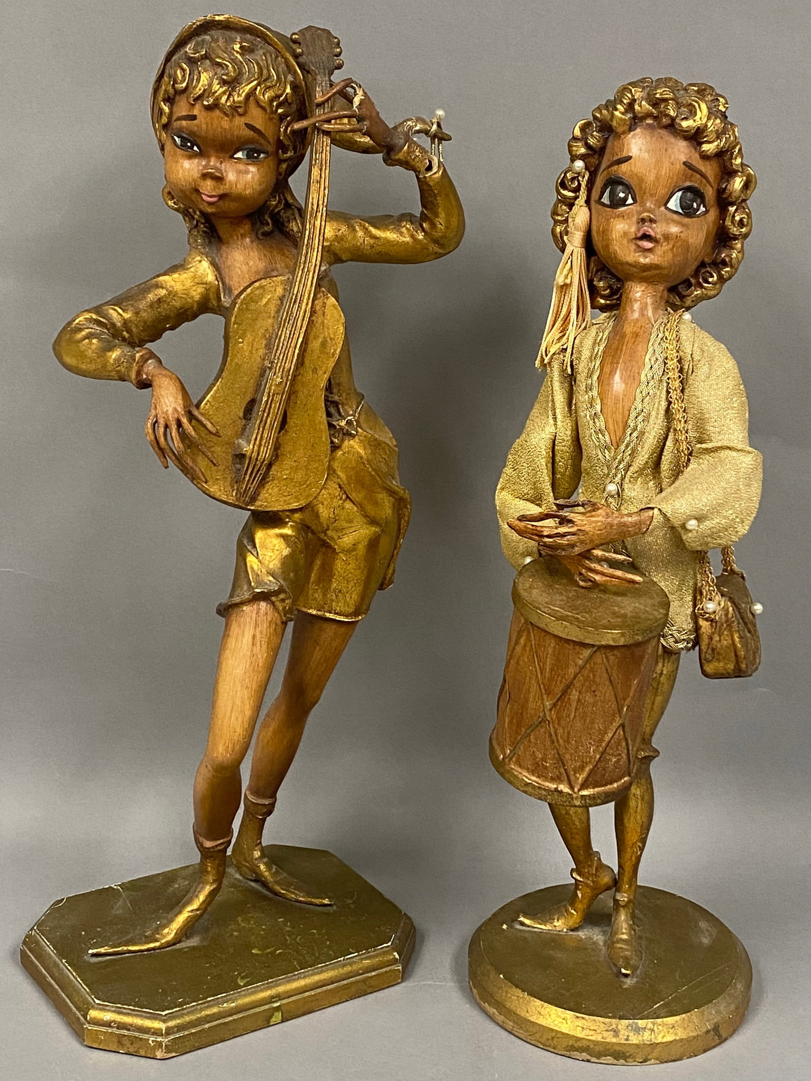 Pair of Mid-Century Musician Figurines: A pair of large-scale mid-century figurines featuring stylized child musicians with oversized eyes and elongated limbs, finished in a wood-grain texture with gilded accents. The girl figure is depicte