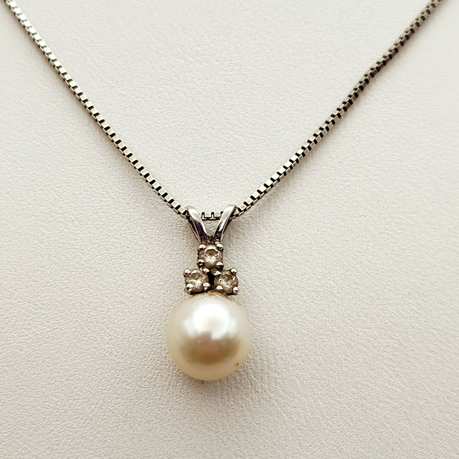 14K Gold Pearl Necklace 3.8 Grams: A round white cultured pearl pendant featuring three round brilliant-cut diamond accents in a triangular setting. The 14 karat white gold pendant is suspended from a matching box link chain. The lobst