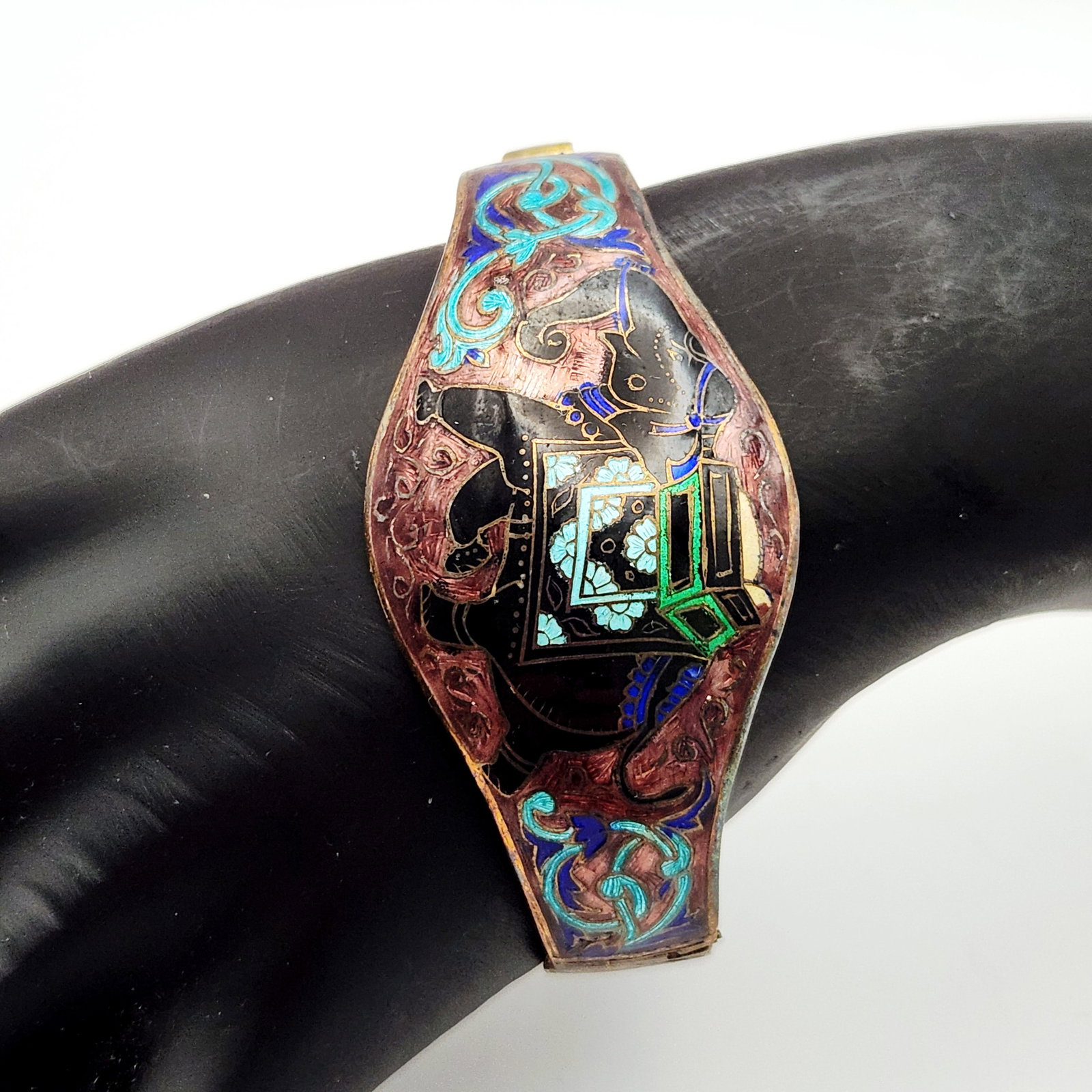 Enamel Elephant Hinged Cuff Bracelet: This hinged cuff bracelet is crafted from darkened yellow metal decorated with champleve-style enamel. The central motif features a caparisoned elephant in black with turquoise and royal blue