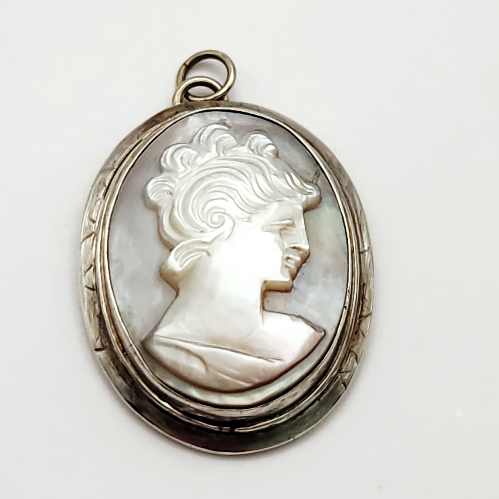 Mother-of-Pearl Cameo Pendant: An oval cameo pendant featuring a hand-carved mother-of-pearl profile of a classical youth set against a dark natural shell backing. The relief is secured in a silver-tone metal bezel frame with a cha
