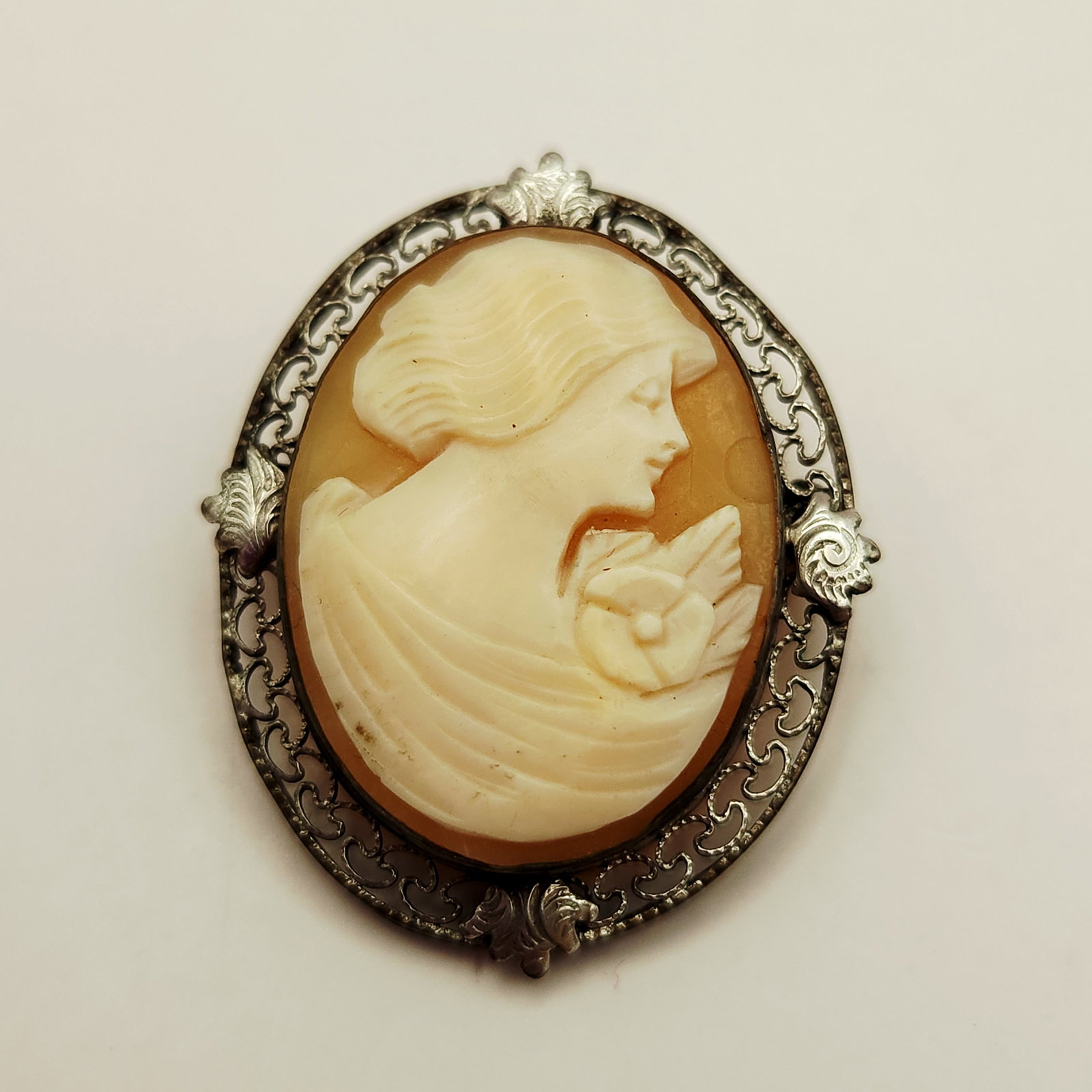 Carved Shell Cameo Filigree Brooch: A mid-20th-century brooch featuring a hand-carved carnelian shell cameo depicting a profile of a woman holding a single flower. The carving is set within an ornate silver-toned metal frame decorated