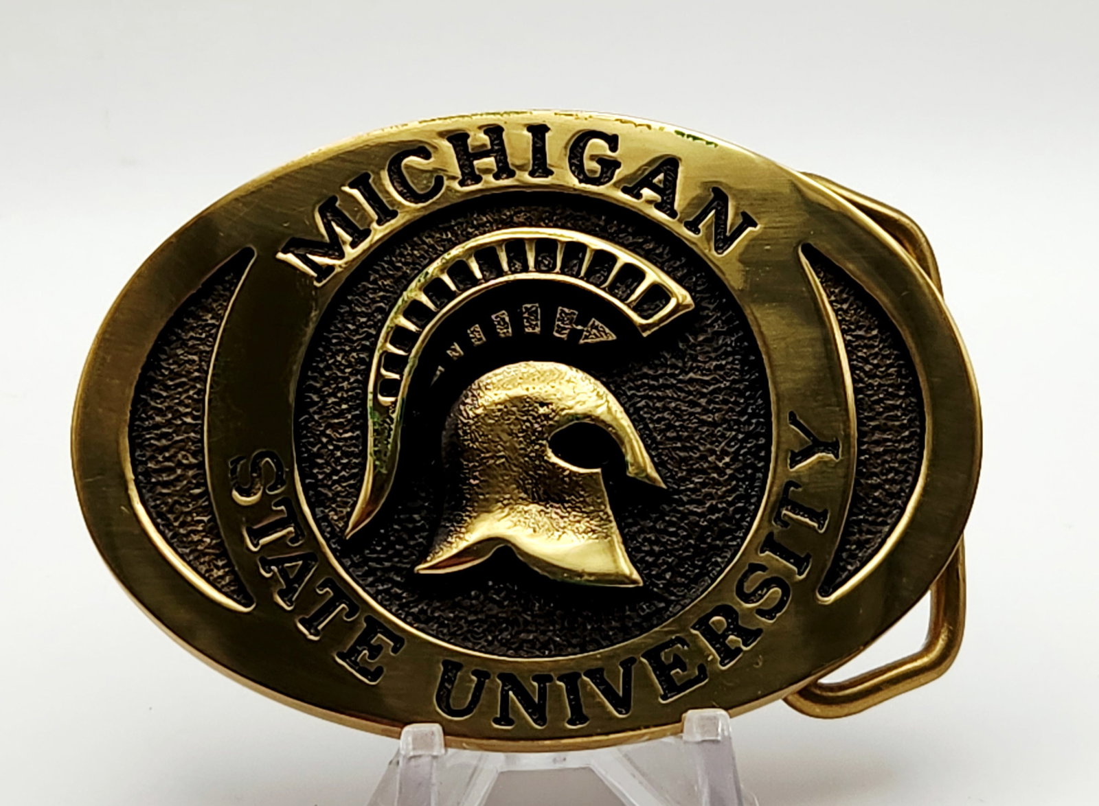 Heritage Mint MSU Brass Buckle: This solid brass oval belt buckle features a high-relief Michigan State University Spartan helmet logo set against a pebbled, dark-patinated ground, serial number EN-3378. The buckle displays minor
