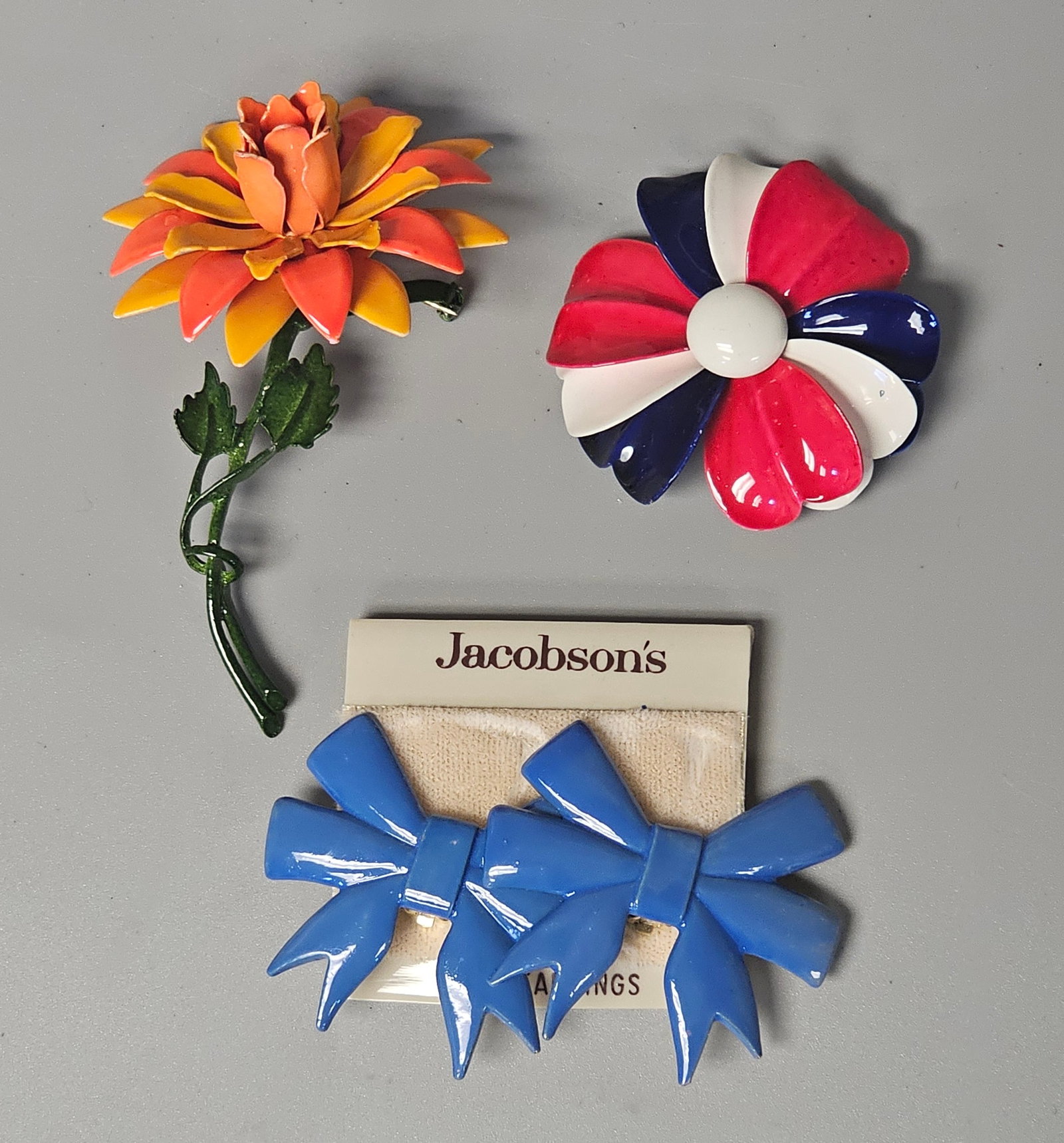 Enameled Floral Brooches and Jacobson's Earrings: This collection of mid-century costume jewelry includes two floral brooches and a pair of clip-on earrings. All pieces are constructed of base metal with glossy enamel finishes. Orange flower brooch: