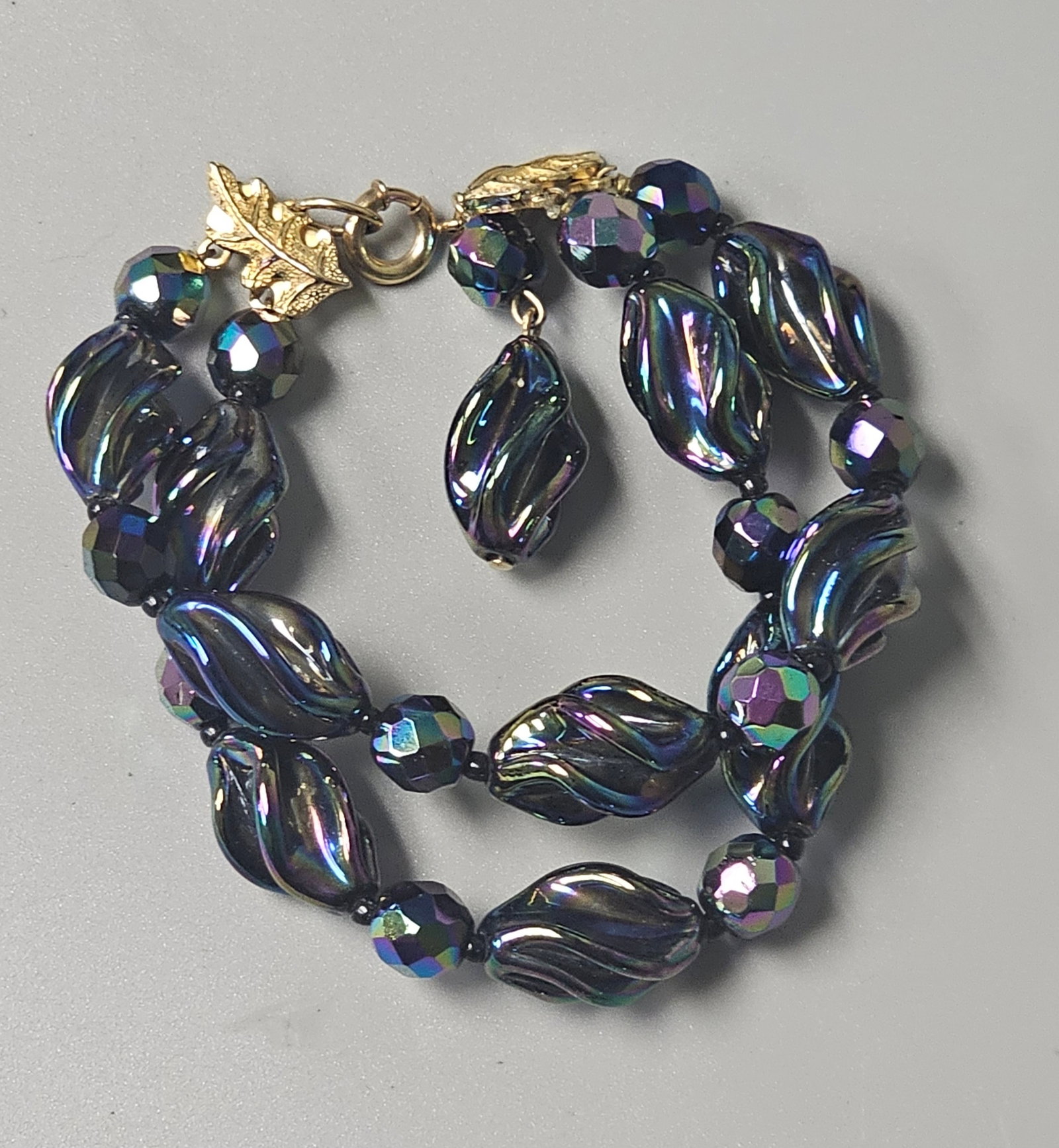Castlecliff Iridescent Glass Two-Strand Bracelet: A vintage mid-century two-strand bracelet featuring glass beads with a vibrant aurora borealis finish in shades of purple, green, and blue. . The reverse of one leaf-shaped connector bears an