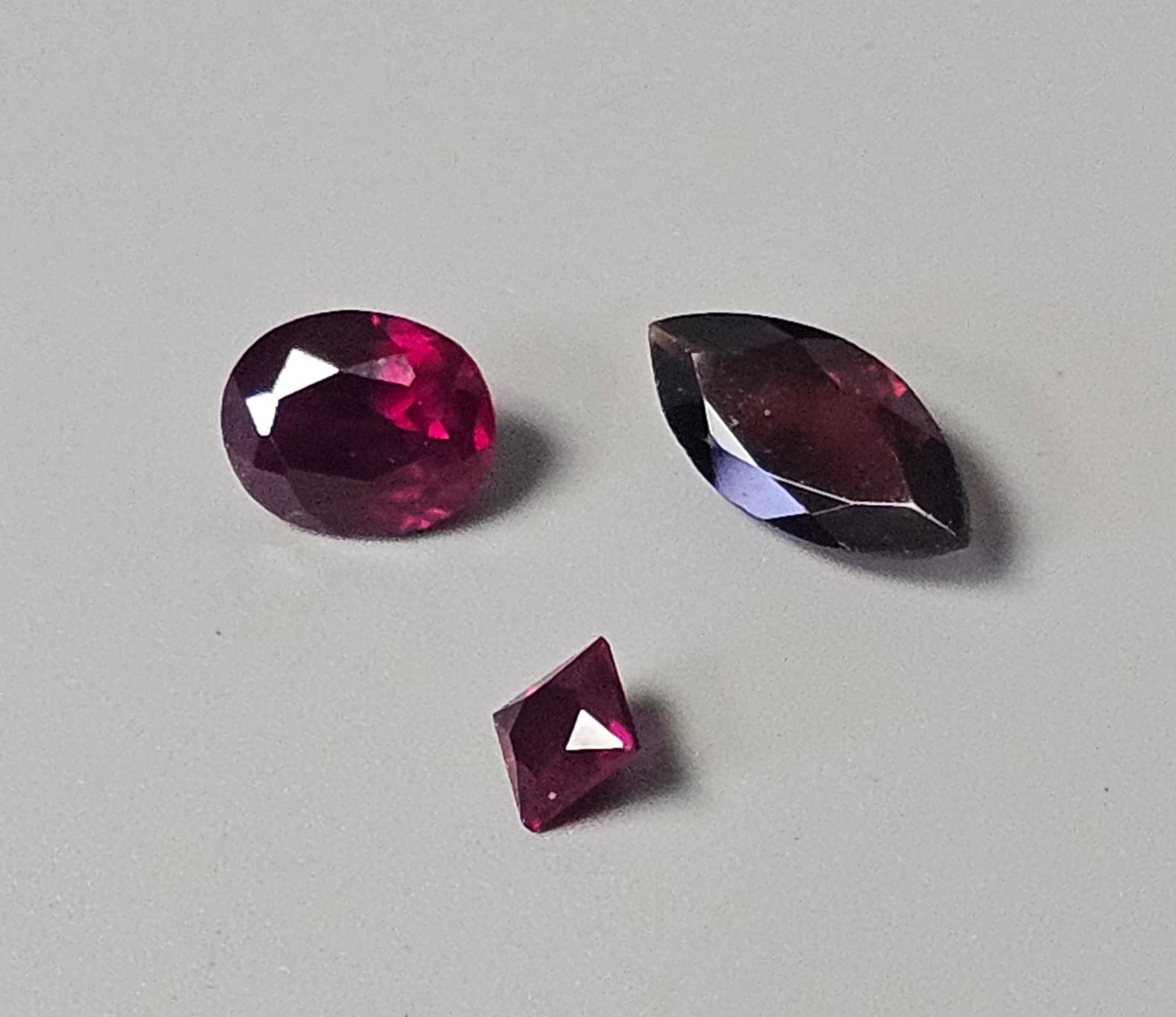 Three Loose Faceted Garnet Gemstones: This lot comprises three loose faceted gemstones identified by the accompanying packaging as garnets. The stones feature oval, marquise, and princess cuts, each displaying a deep, saturated