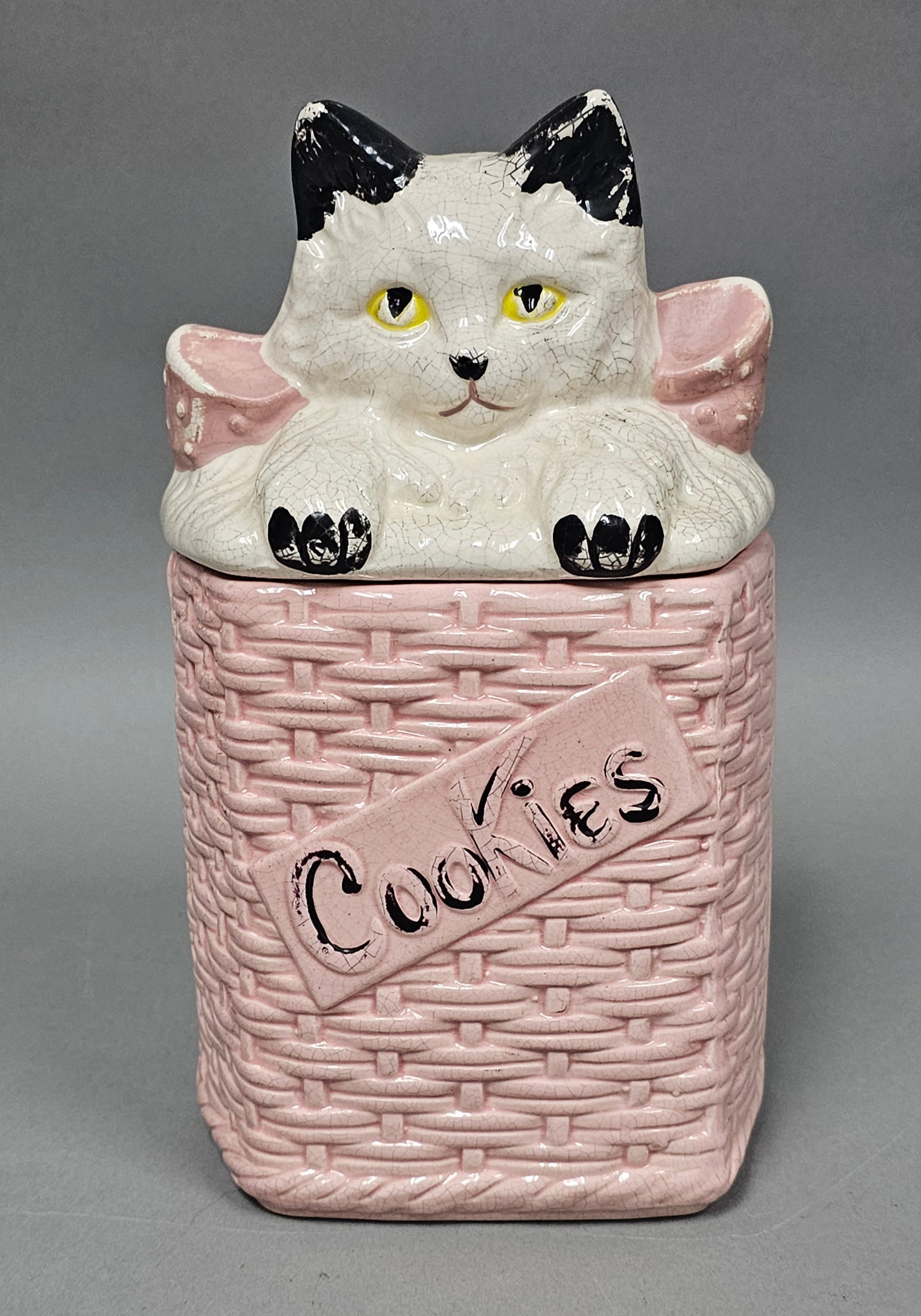 McCoy Ceramic Kitty Cat Basket Cookie Jar: A vintage earthenware cookie jar manufactured by McCoy Pottery. The piece features a rectangular base molded with a textured basket-weave pattern and a diagonal plaque on the front inscribed with the 