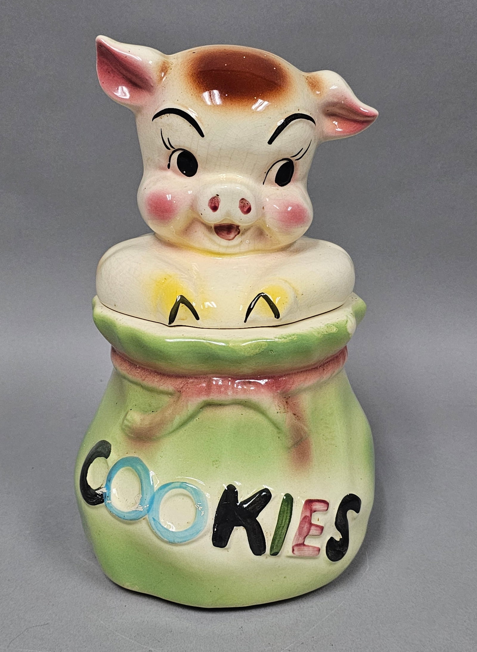 USA Ceramic Pig in Sack Cookie Jar: This vintage figural cookie jar is crafted from glazed earthenware in two pieces. The base is shaped as a gathered green sack with a pink rope tie, featuring the word COOKIES in relief block letters o