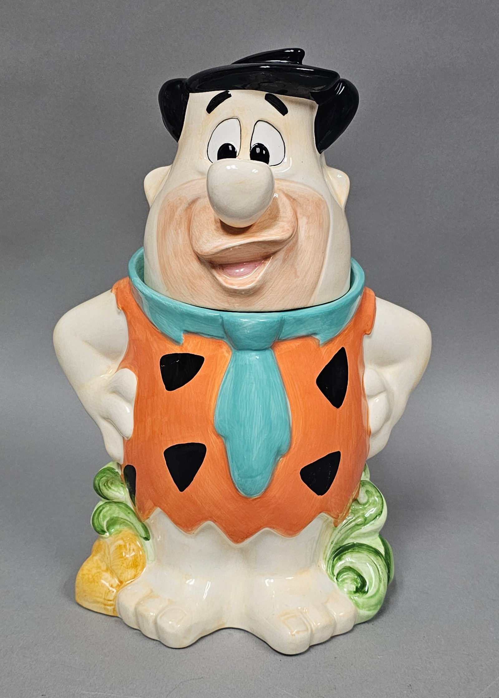 Certified International Fred Flintstone Cookie Jar: This two-piece ceramic cookie jar depicts the Hanna-Barbera character Fred Flintstone from The Flintstones. The figure is posed in a classic stance with hands on hips, wearing an orange leopard-print