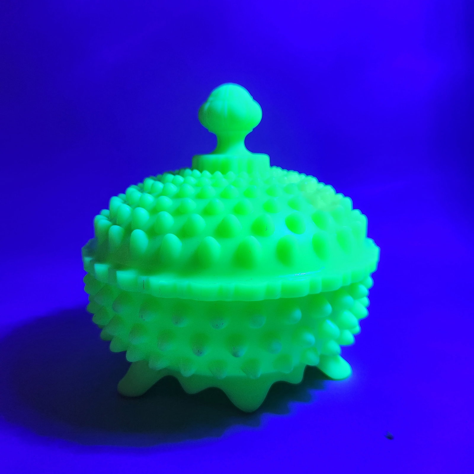Fenton Custard Hobnail Candy Dish: This lidded candy dish is crafted in Fenton uranium custard glass featuring the pressed hobnail pattern. The three-footed bowl and matching lid exhibit a strong green fluorescence under ultraviolet