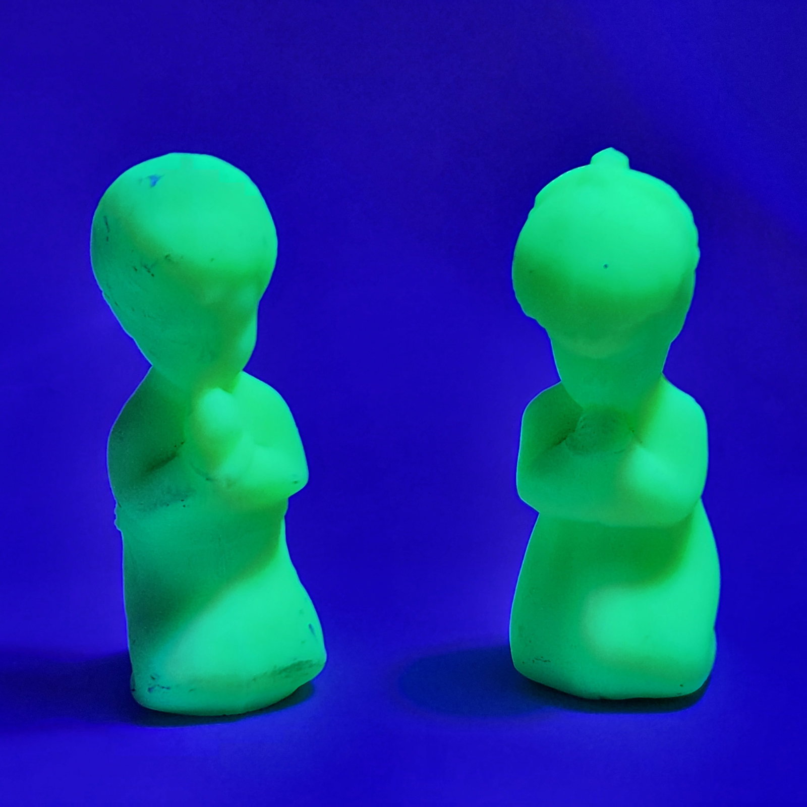 Fenton Uranium Glass Praying Children: A matched pair of Fenton custard glass figurines depicting a young boy and girl in prayer. The opaque light green glass displays a strong green fluorescence under ultraviolet light, confirming the