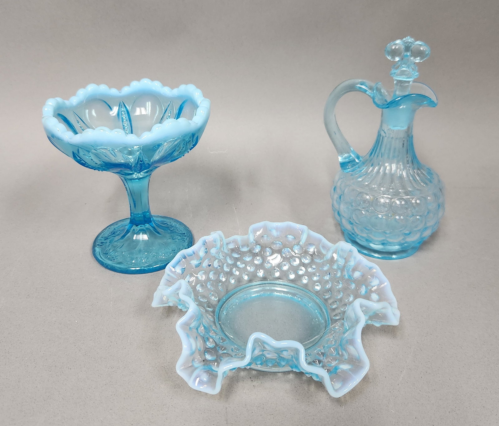 Trio of Fenton Blue Glass: This lot features three Fenton glass articles in varying shades of blue, including a pedestal compote, a ruffled bowl, and a cruet with a matching stopper. The pieces showcase classic opalescent and