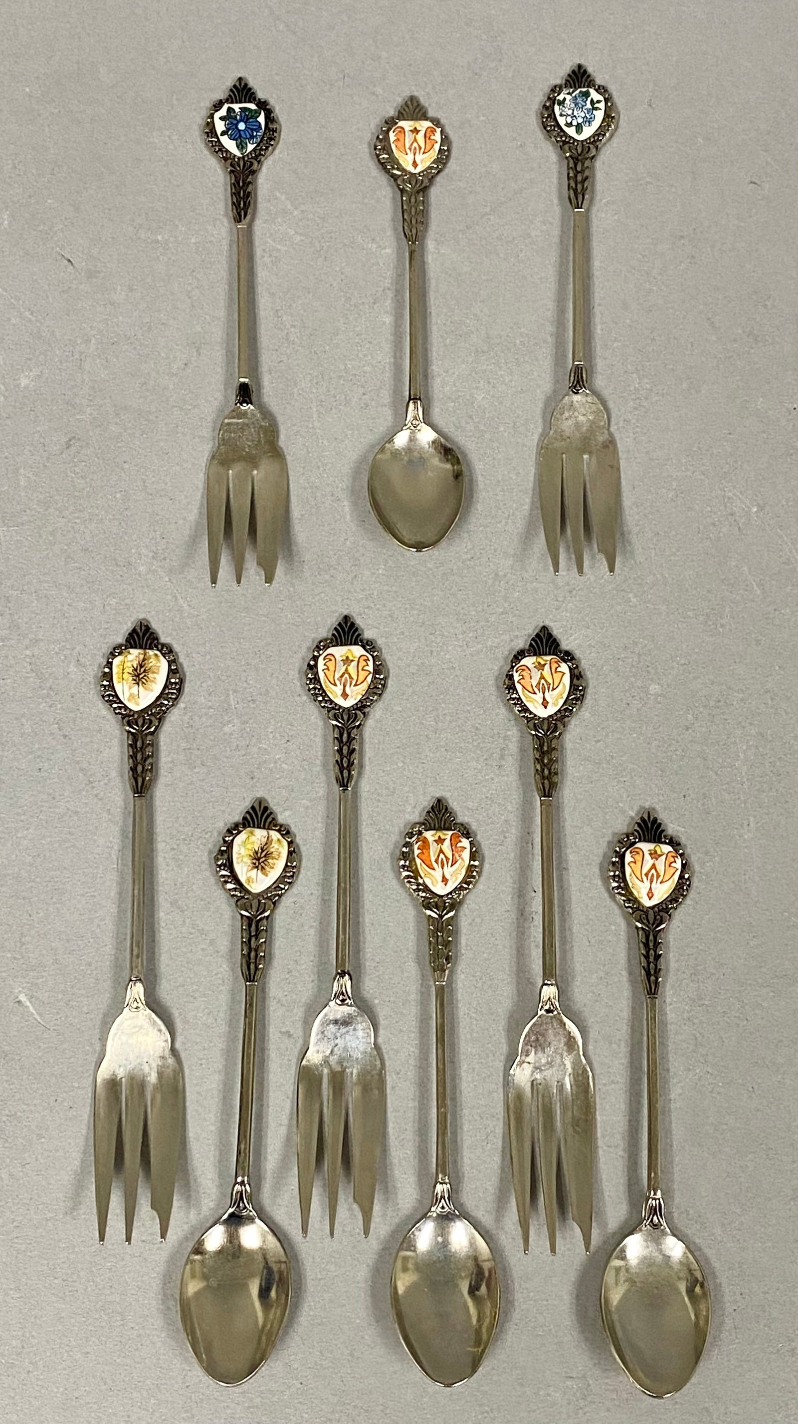 9pc Japanese Silver-Tone Enamel Dessert Flatware: A nine-piece set of Japanese silver-tone metal dessert flatware consisting of five forks and four spoons. The handles are designed with ornate openwork scrollwork and terminate in shield-shaped