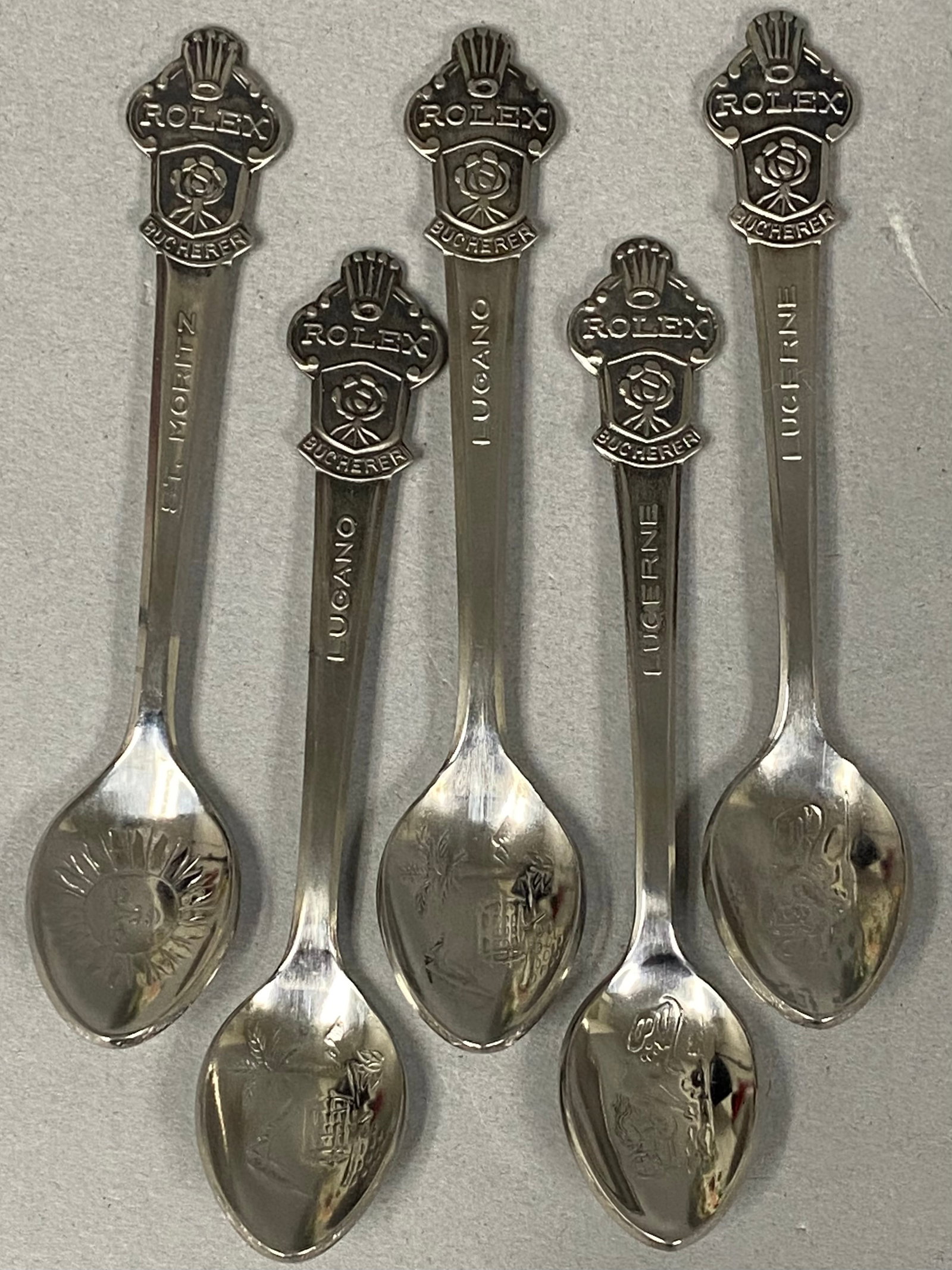 5 Rolex Bucherer Silver-Plated Souvenir Spoons: A set of five silver-plated souvenir spoons commissioned by Rolex for Bucherer. Each demitasse-sized spoon features a decorative finial topped with the Rolex coronet logo above the name ROLEX, a
