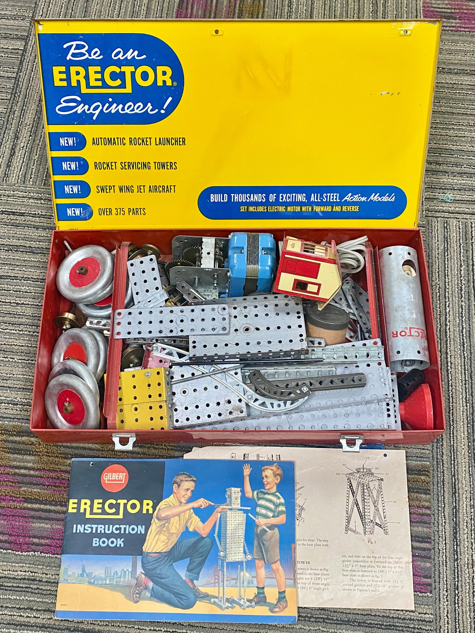 Gilbert Steel No. 10063 Action Conveyor Erector Set: An assortment of perforated steel plates, girders, and mechanical components from a Gilbert No. 10063 Action Conveyor Erector Set. The collection is housed in its original red and yellow lithographed