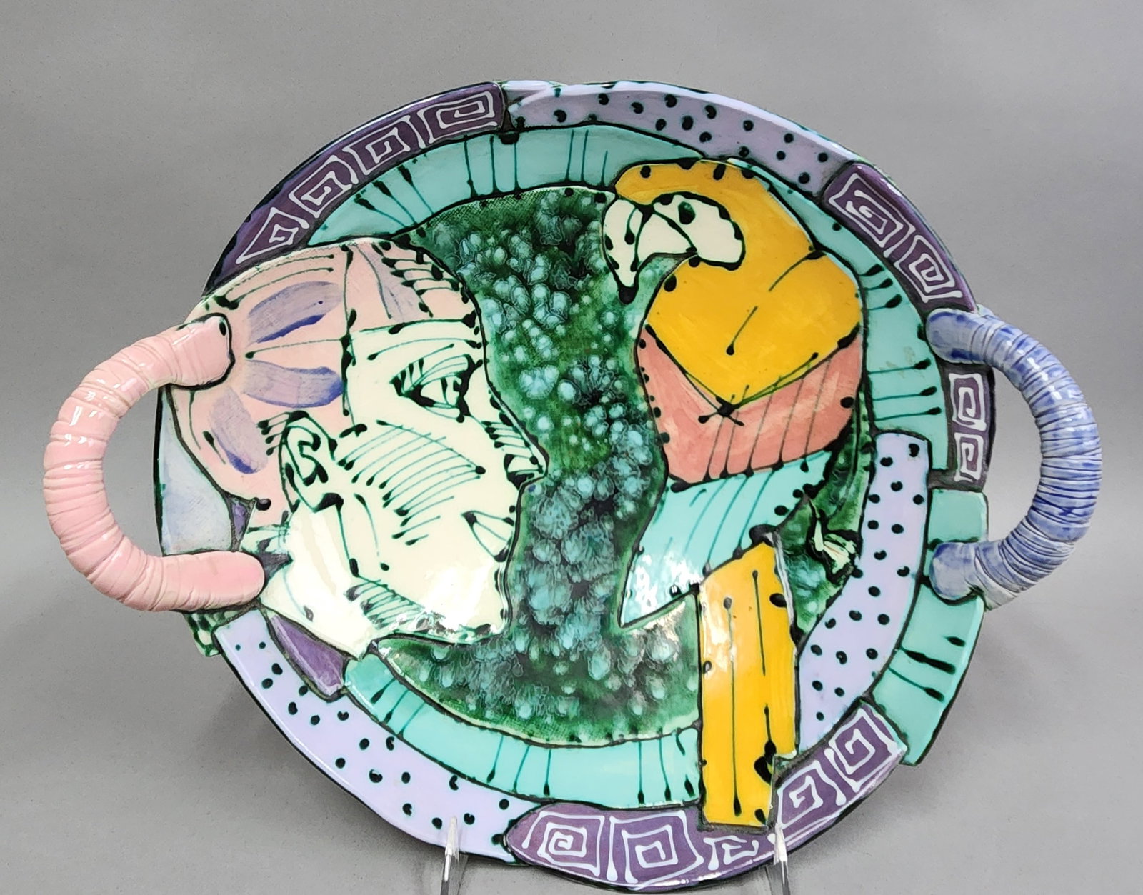 Ceramic Figurative Handled Bowl: This hand-painted contemporary studio ceramic bowl features a shallow circular form with two oversized, annulated C-scroll handles finished in pastel pink and periwinkle blue. In the style of