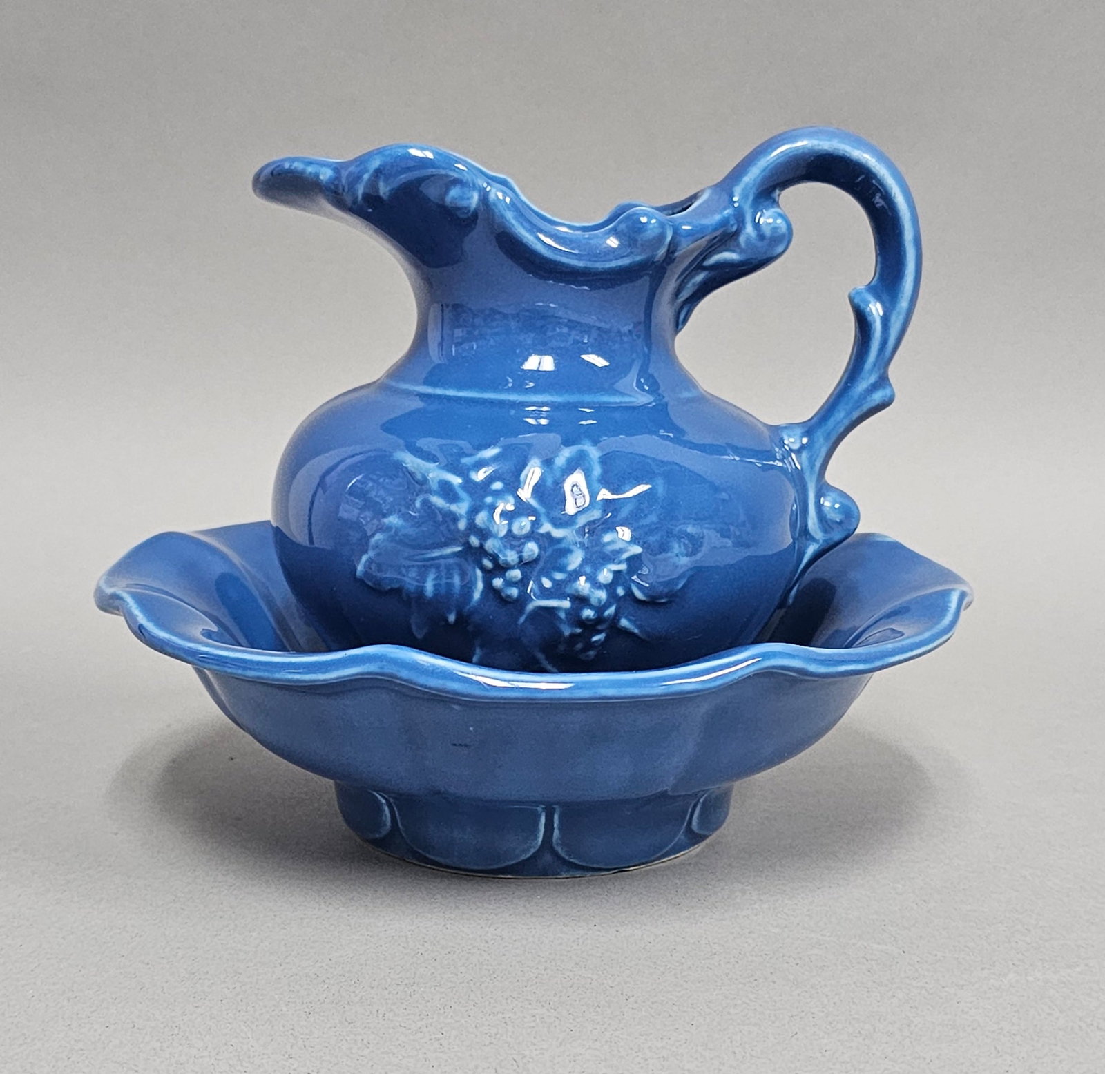 McCoy Ceramic Blue Grape Pitcher & Basin Set: A ceramic miniature wash pitcher and basin set from McCoy Pottery finished in a vibrant, glossy royal blue monochrome glaze. The bulbous pitcher is decorated with a molded relief of a grape cluster an