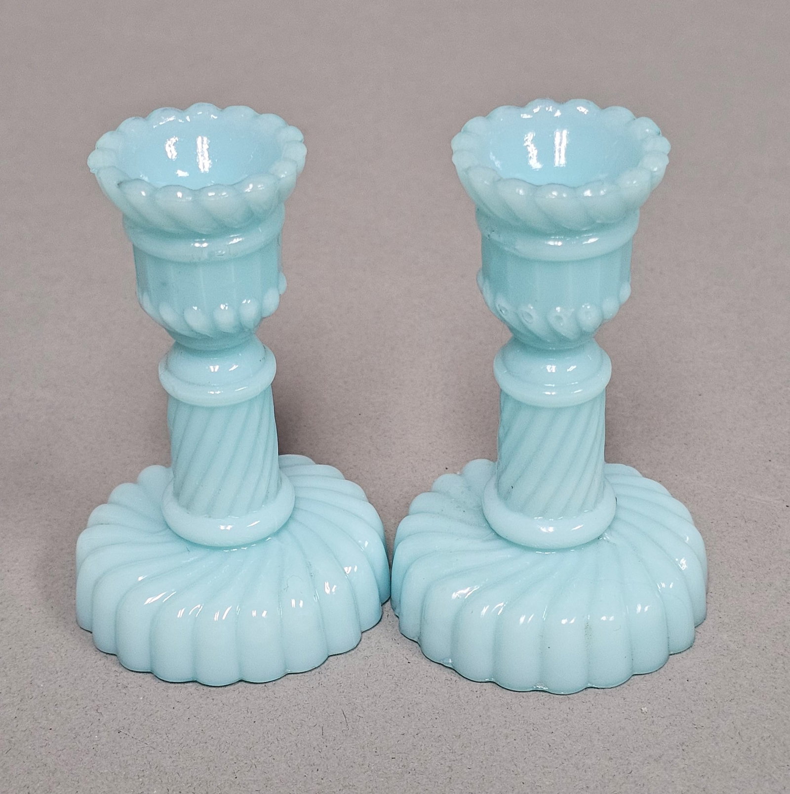 Blue Milk Glass Spiral Candlesticks: A pair of matching pressed glass candlesticks crafted from opaque light blue milk glass. Each piece features a spiral-ribbed stem set upon a wide circular base with gadrooned ribbing and a scalloped e
