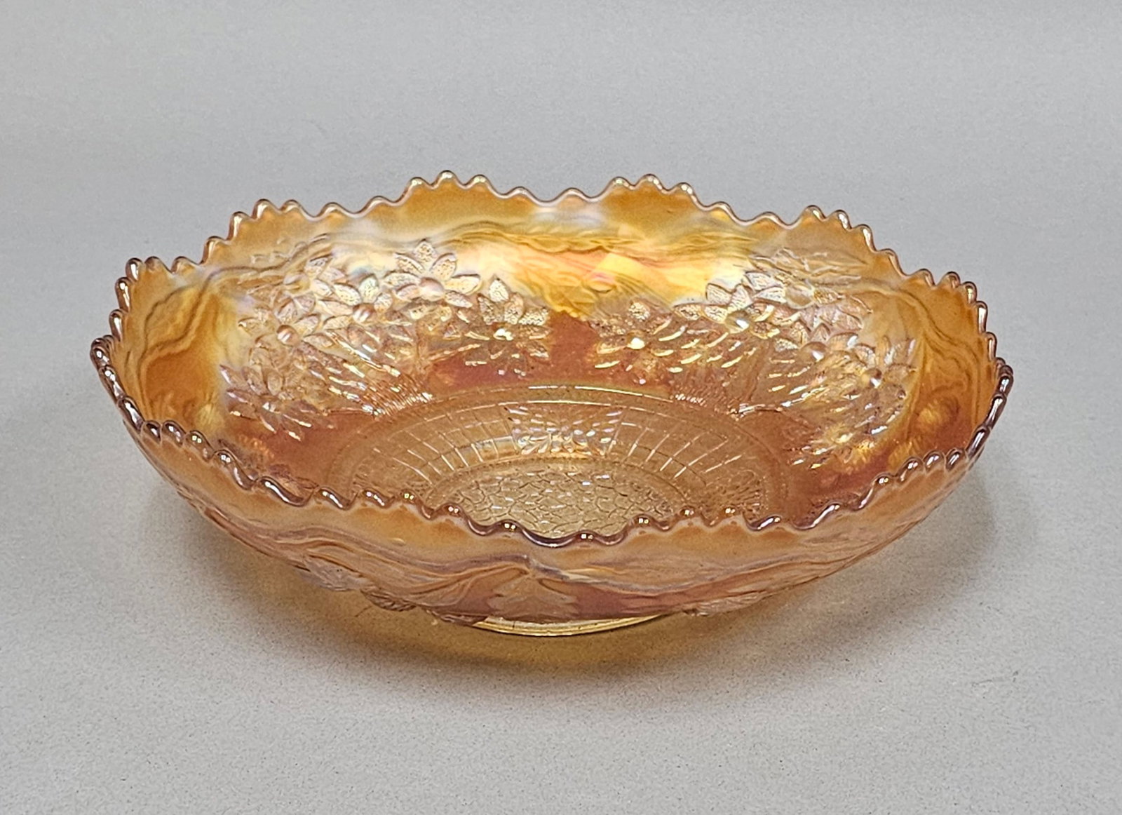 Marigold Carnival Glass Ruffled Bowl: A pressed iridescent glass bowl in a marigold color featuring a wide ruffled rim with a sawtooth edge. The interior is decorated with a molded floral design featuring a central circular medallion