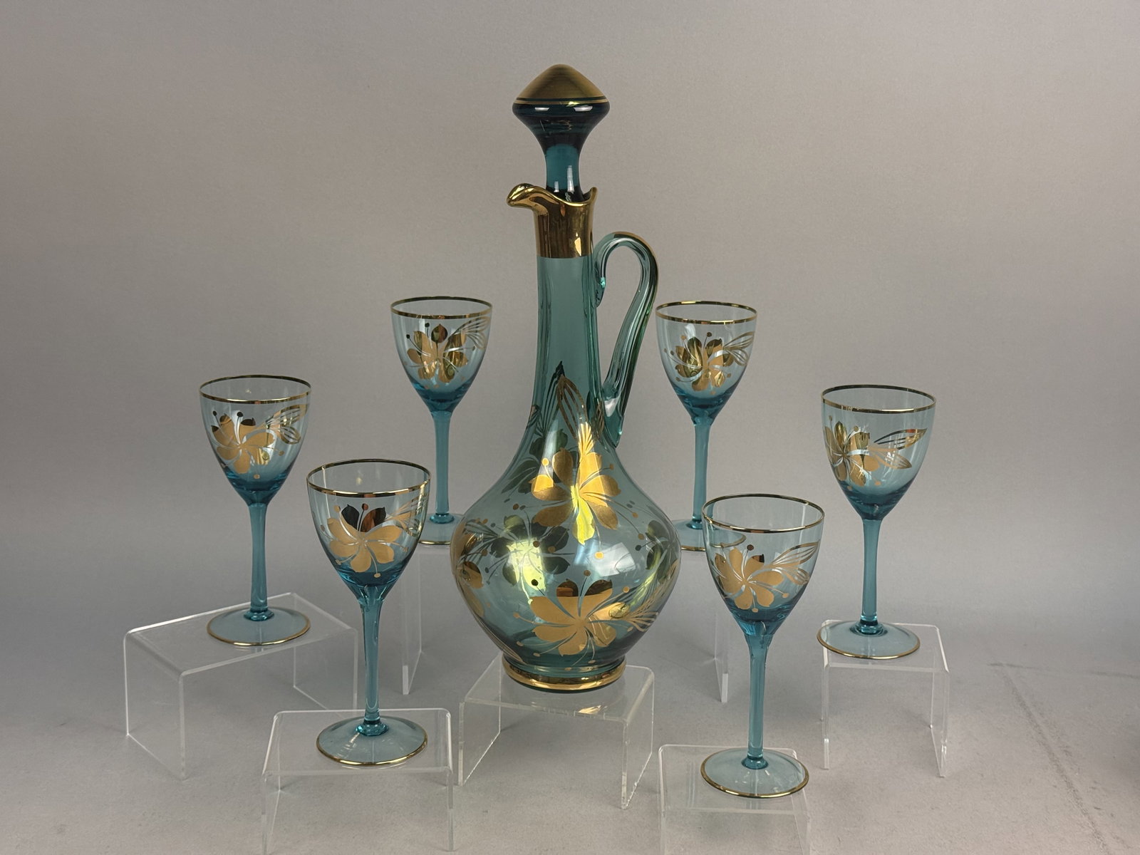 Romanian Teal and Gilt Glass Set: This Romanian mid-century modern beverage set is crafted from hand-blown teal glass featuring hand-painted gold floral motifs and black enamel accents. The set includes a handled decanter with a match