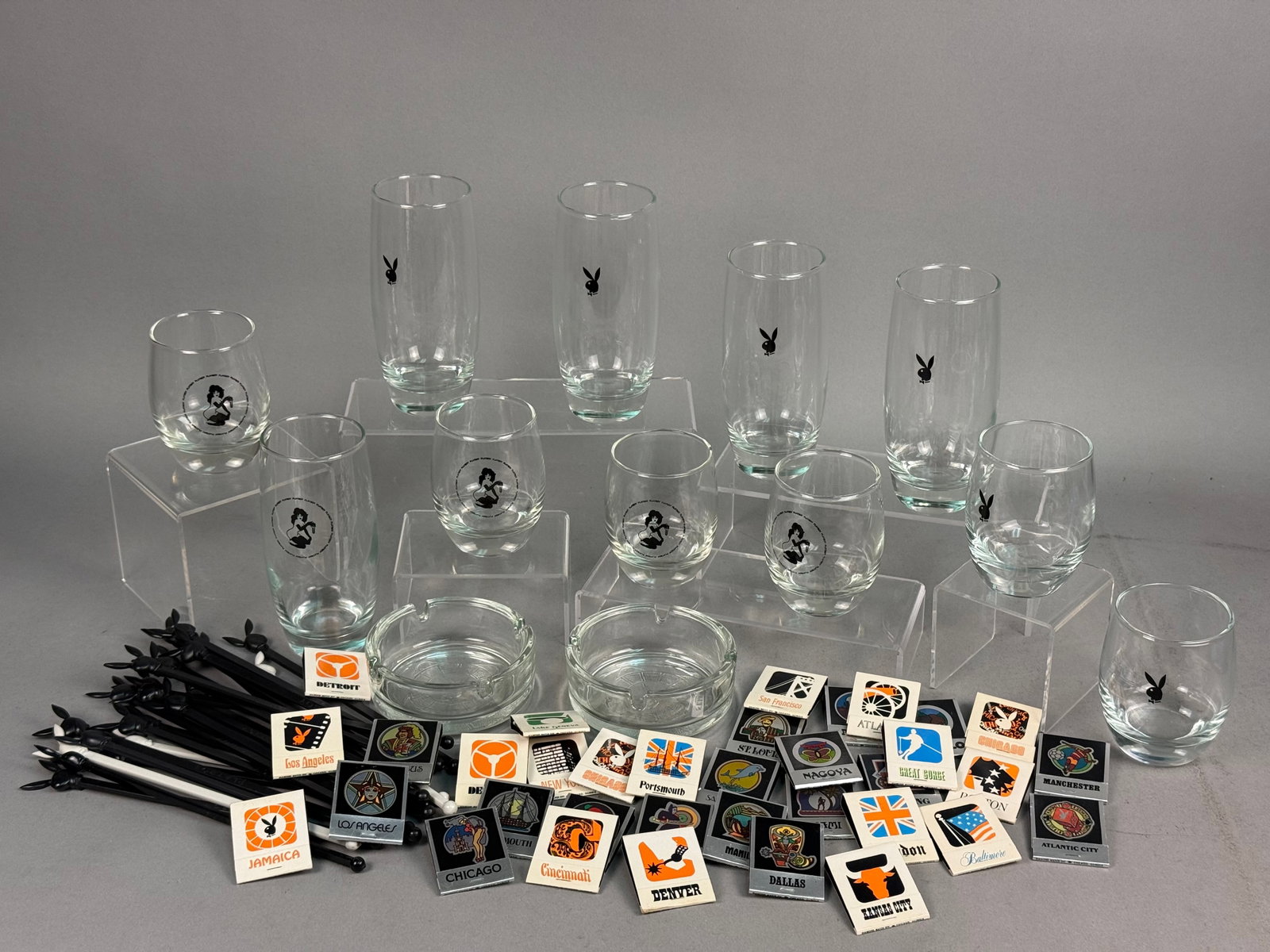 Playboy Glass Barware & Memorabilia: An assortment of vintage barware and promotional materials from the Playboy Club. The set includes clear glass highballs featuring a black printed bunny head logo. Also present are clear glass rocks