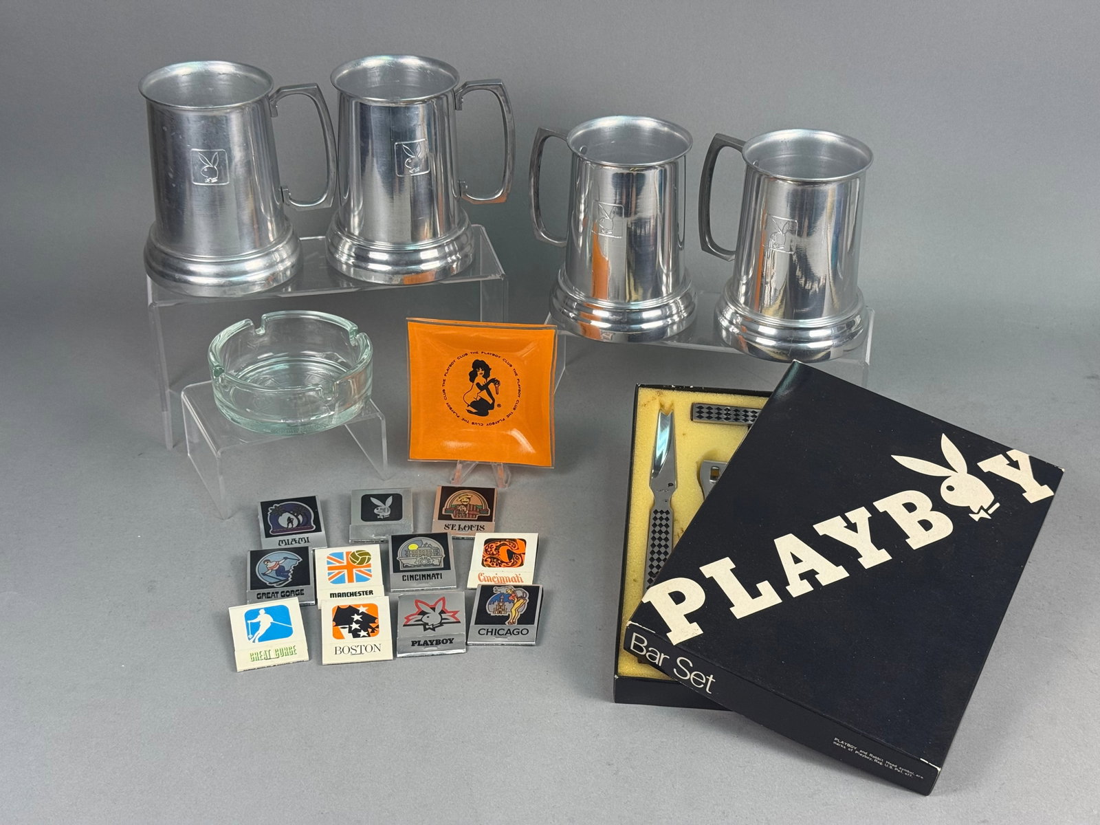Playboy Aluminum & Glass Barware Collection: An assortment of mid-century Playboy themed barware featuring four aluminum tankards with clear glass bottoms. Each mug displays an embossed rabbit head logo within a square cartouche. The set include