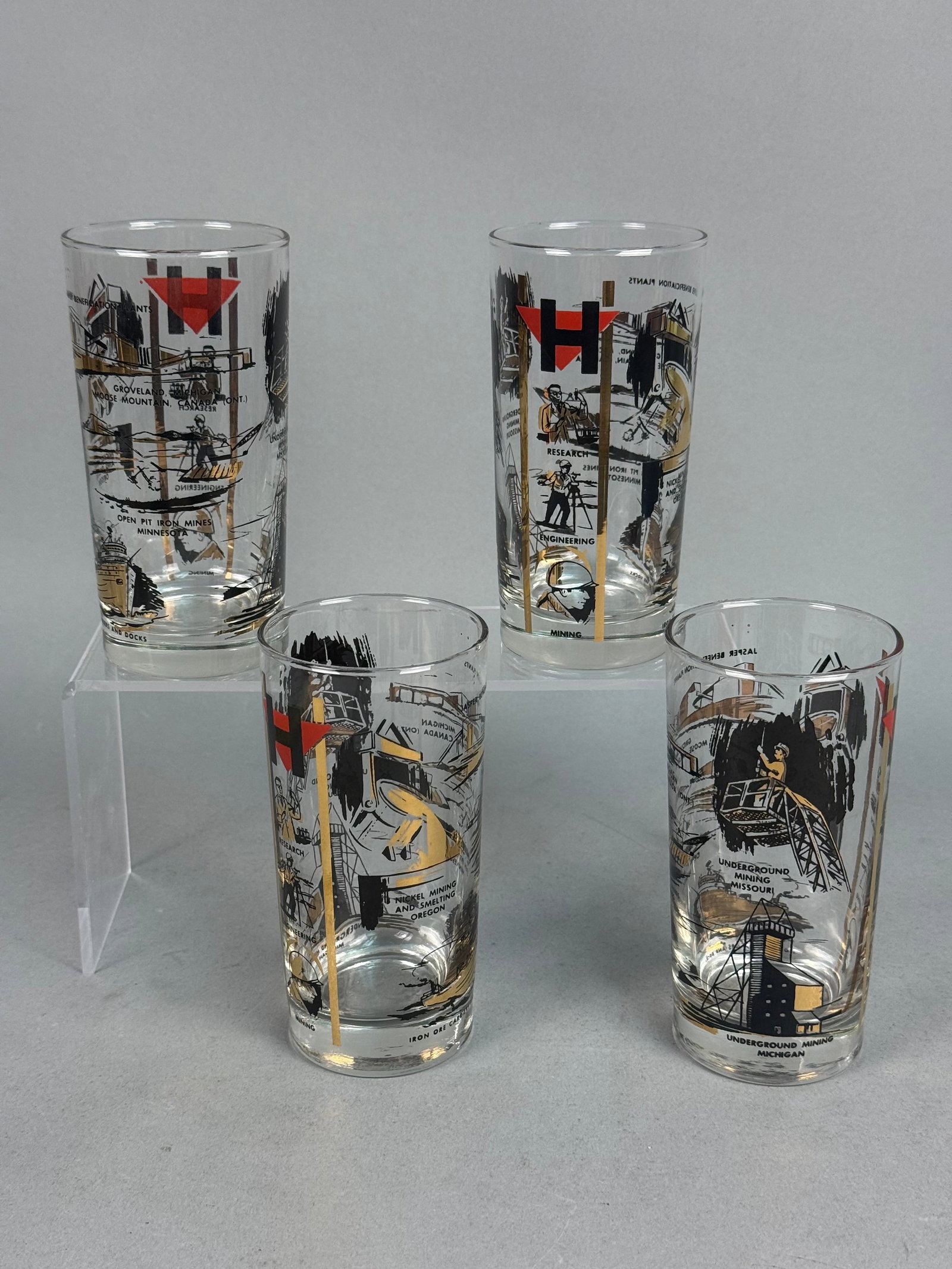 Hanna Mining Industrial Glass Tumblers: A set of four clear glass tumblers featuring wrap-around industrial graphics documenting the operations of the Hanna Mining Company. The designs are executed in black and red fired-on enamel,